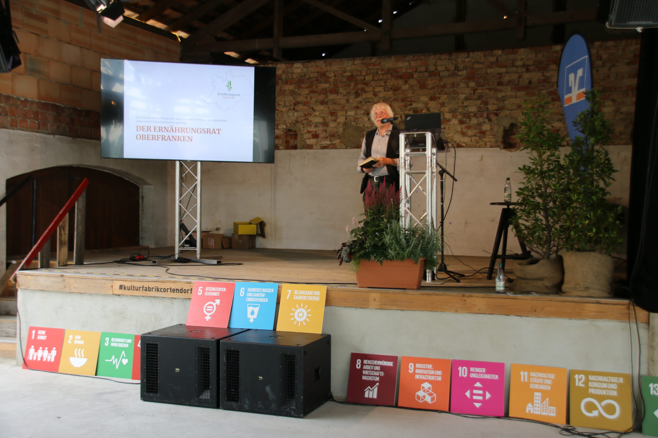 A person stands on a small stage at Hochschule Coburg with a microphone, presenting near a large screen that reads "Der Ernährungsrat Oberfranken." Various colorful sustainable development goal icons are displayed in front of the stage.