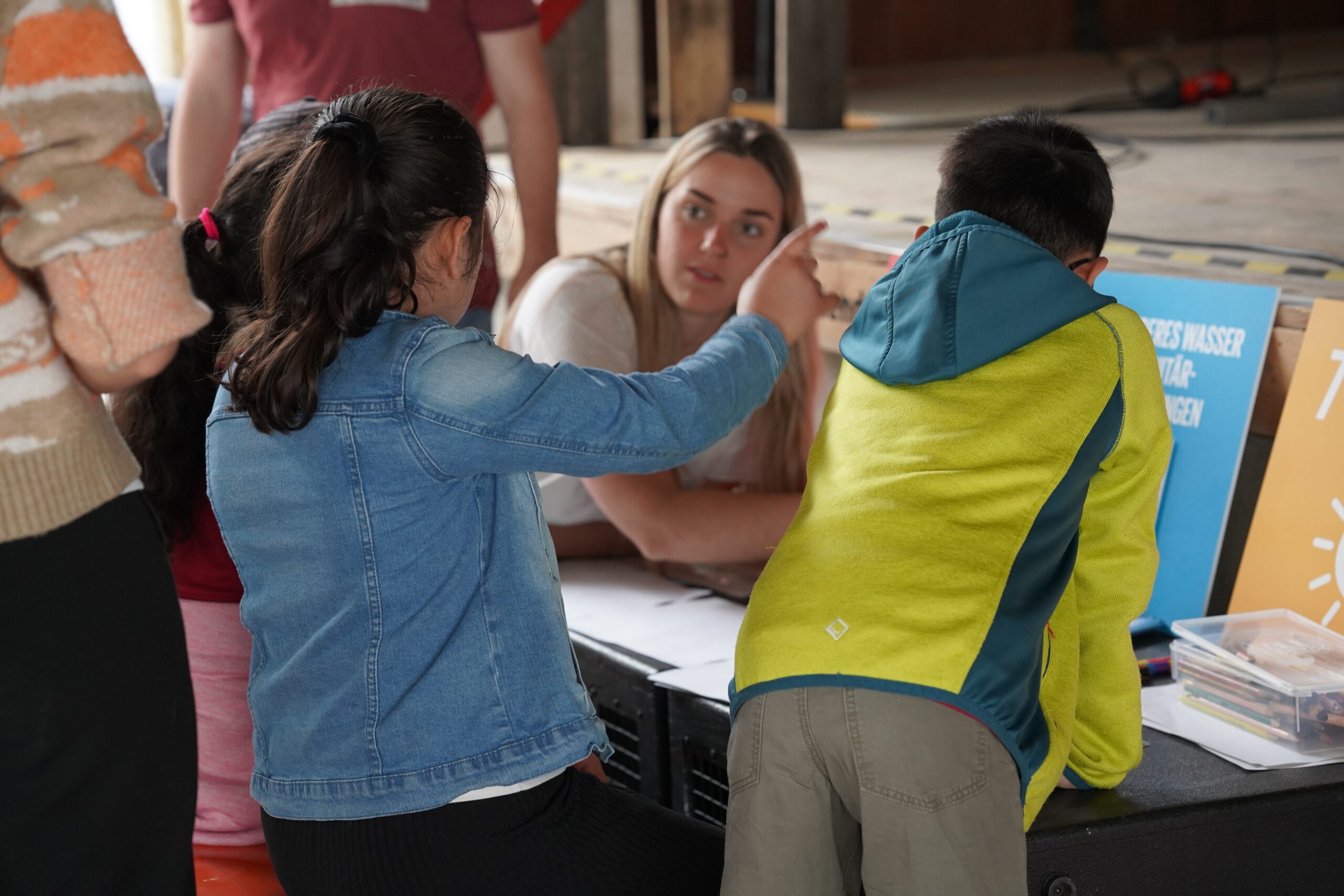 A group of young people is interacting around a table at Hochschule Coburg. A girl in a denim jacket is gesturing towards a boy in a green and yellow jacket. A woman with long hair sits behind them, observing papers and a sign on the table.