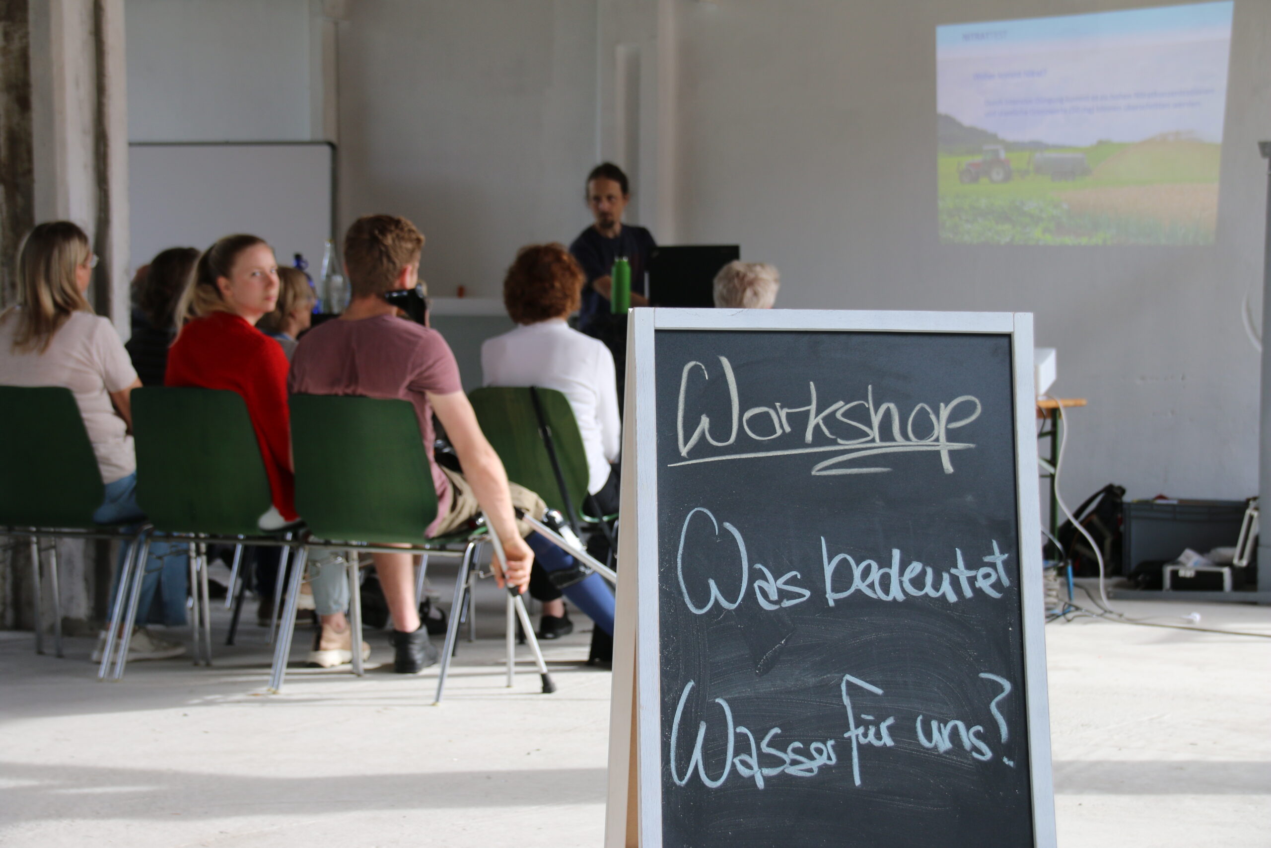 In einem hellen Raum der Hochschule Coburg sitzt eine Gruppe auf grünen Stühlen einem Vortragenden gegenüber. Neben der Leinwand, auf der eine Landschaft zu sehen ist, steht auf einem Schild: „Workshop: Was bedeutet Wasser für uns?“