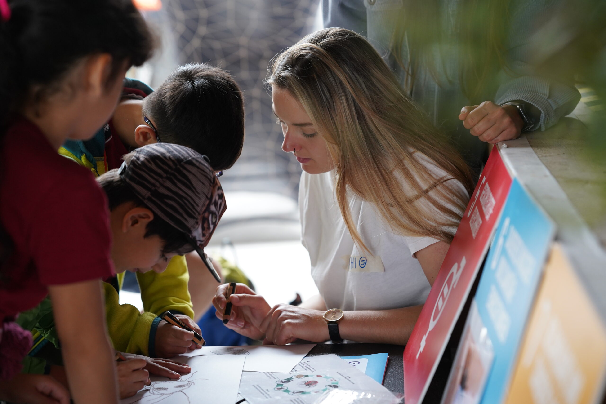 A woman with long blonde hair, perhaps an alumna of Hochschule Coburg, helps a group of children with an art project. The children, dressed casually, focus intently on drawing, creating a lively and colorful scene that suggests a creative and interactive environment.