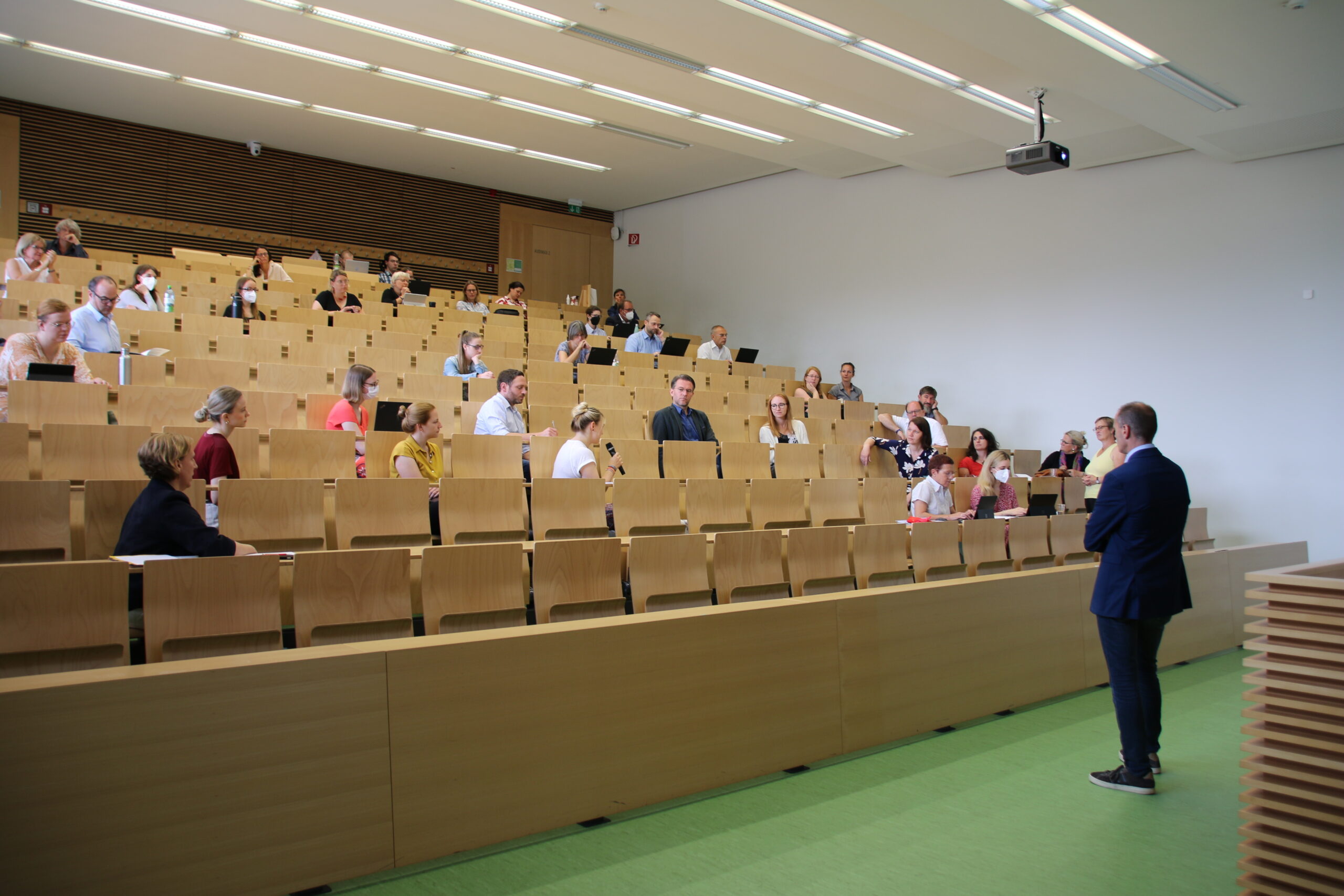 Ein Dozent steht vor einem Hörsaal der Hochschule Coburg und spricht zu einem Publikum, das in Reihen von Holzbänken sitzt. Die Teilnehmer sind über den geräumigen Raum verteilt und hören aufmerksam zu.