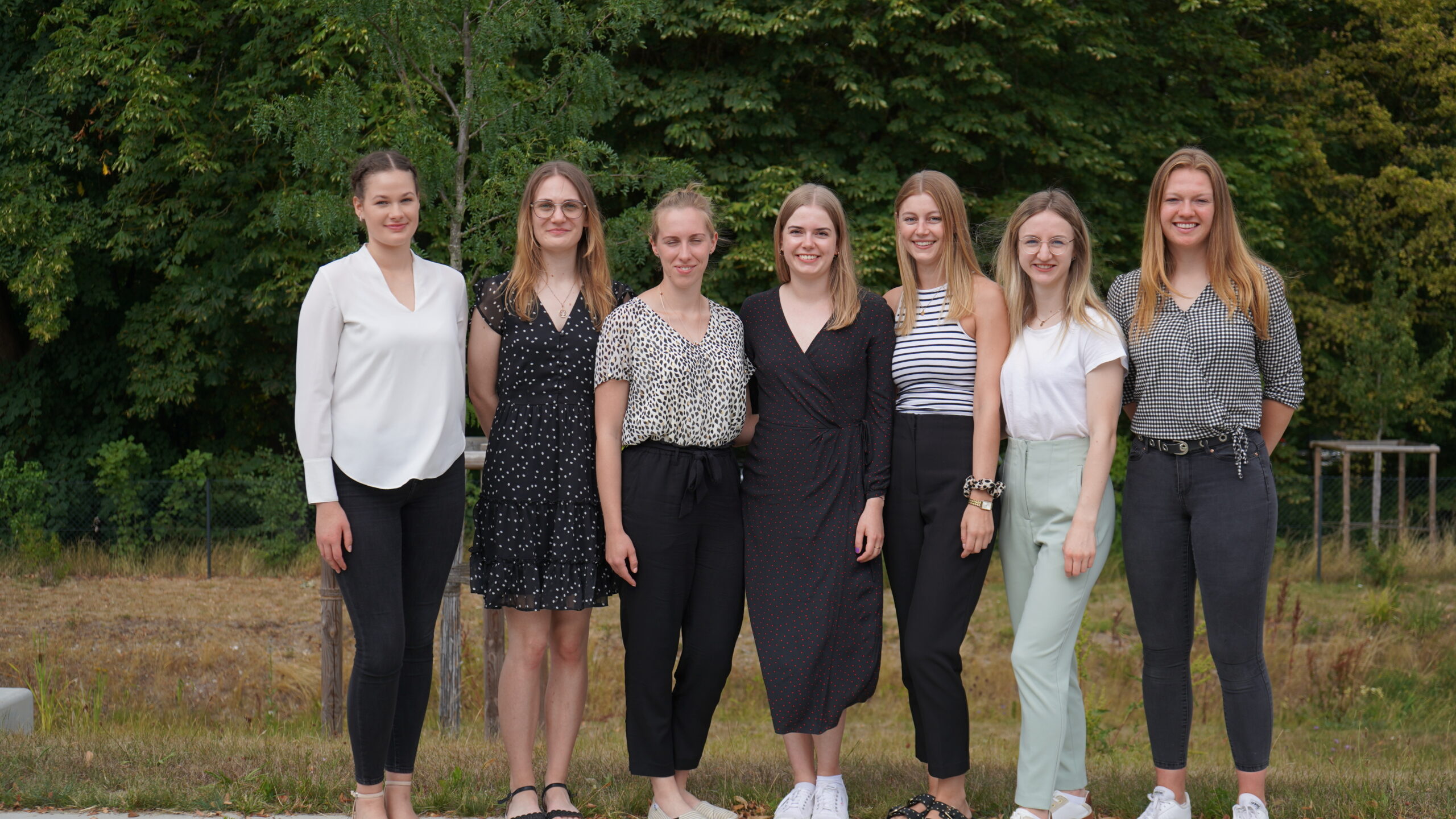 Sieben Frauen stehen in einer Reihe im Freien und lächeln in die Kamera. In lässiger, aber dennoch stilvoller Kleidung posieren sie stolz vor einer Kulisse aus grünen Bäumen und Gras und repräsentieren die Hochschule Coburg mit Anmut und Kameradschaft.