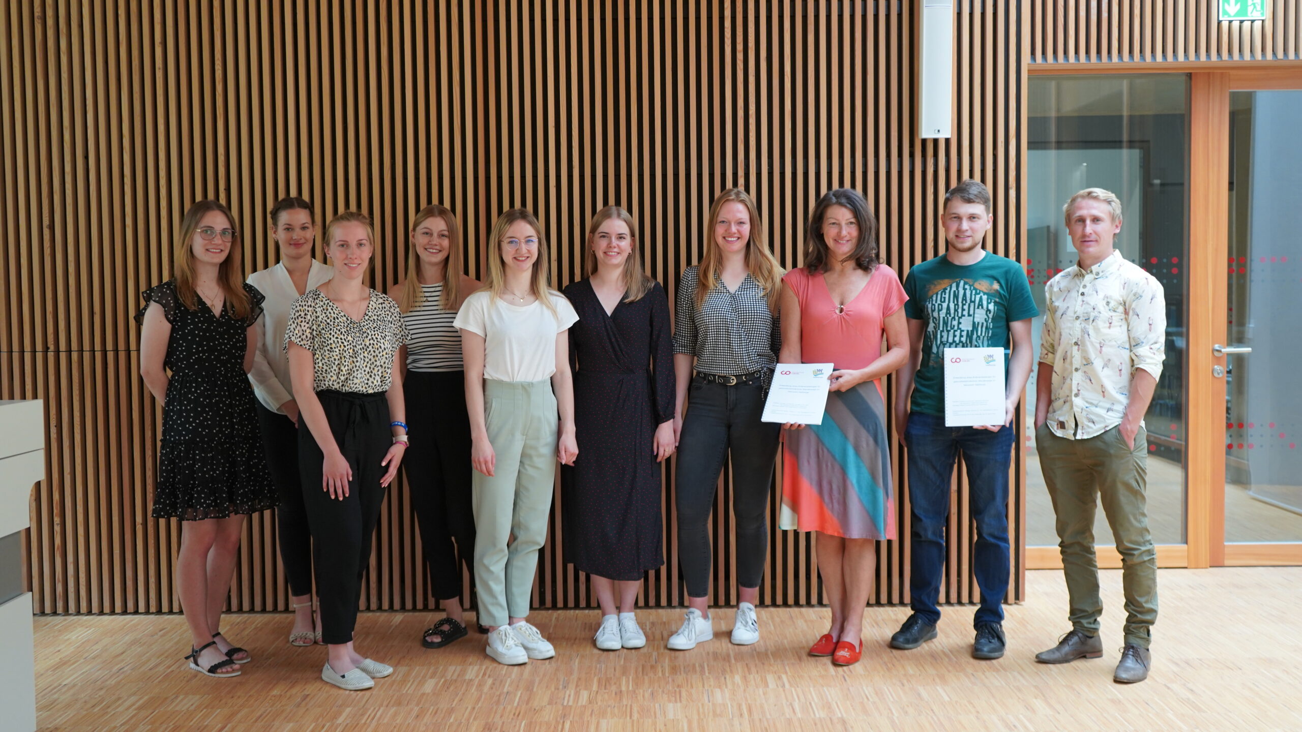 Eine Gruppe von elf Personen steht in einer Reihe in einem Innenbereich der Hochschule Coburg und lächelt stolz. Zwei Personen halten Zertifikate in den Händen und zeigen ihre Leistungen. Im Hintergrund sind Holzvertäfelungen und eine Glastür zu sehen, die dem feierlichen Moment einen Hauch von Eleganz verleihen.