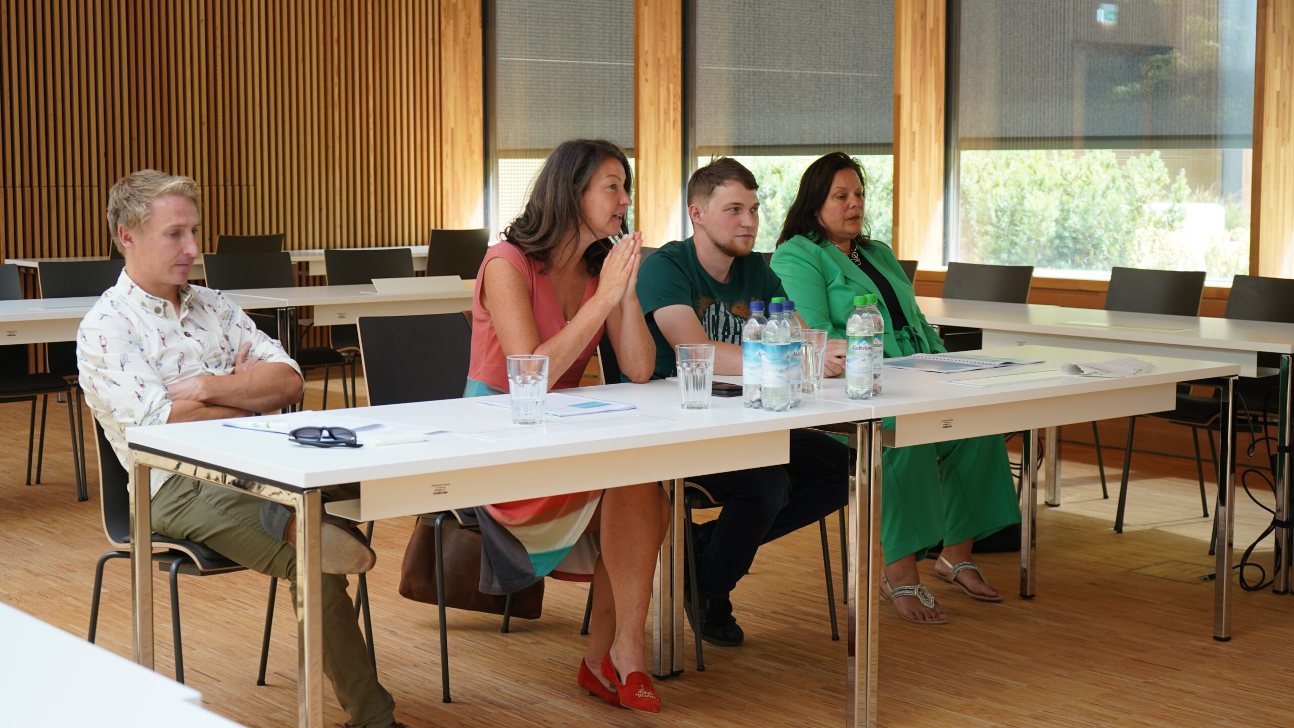 Vier Personen sitzen an einem langen Tisch in einem Konferenzraum mit Holzwänden der Hochschule Coburg und hören aufmerksam zu. Vor ihnen liegen Papiere, Wasserflaschen und Gläser. Eine Person spricht in ein Mikrofon.