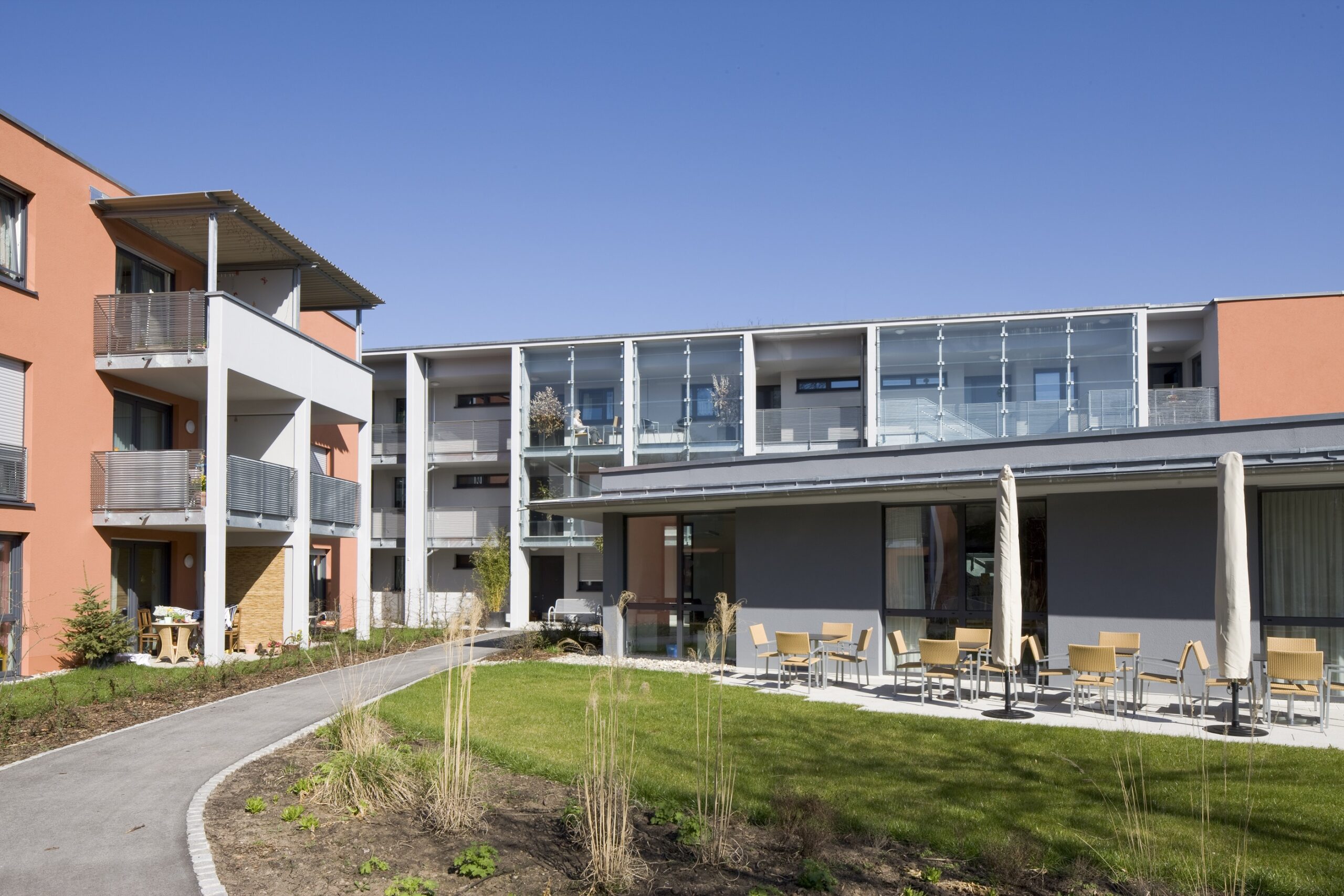 Ein modernes Wohngebäude in der Nähe der Hochschule Coburg verfügt über mehrere Balkone, große Fenster und einen Weg, der sich durch eine Rasen- und Landschaftsgartenfläche schlängelt. Auf einer Terrasse sind Sitzgelegenheiten im Freien unter dem klaren, blauen Himmel zu sehen.