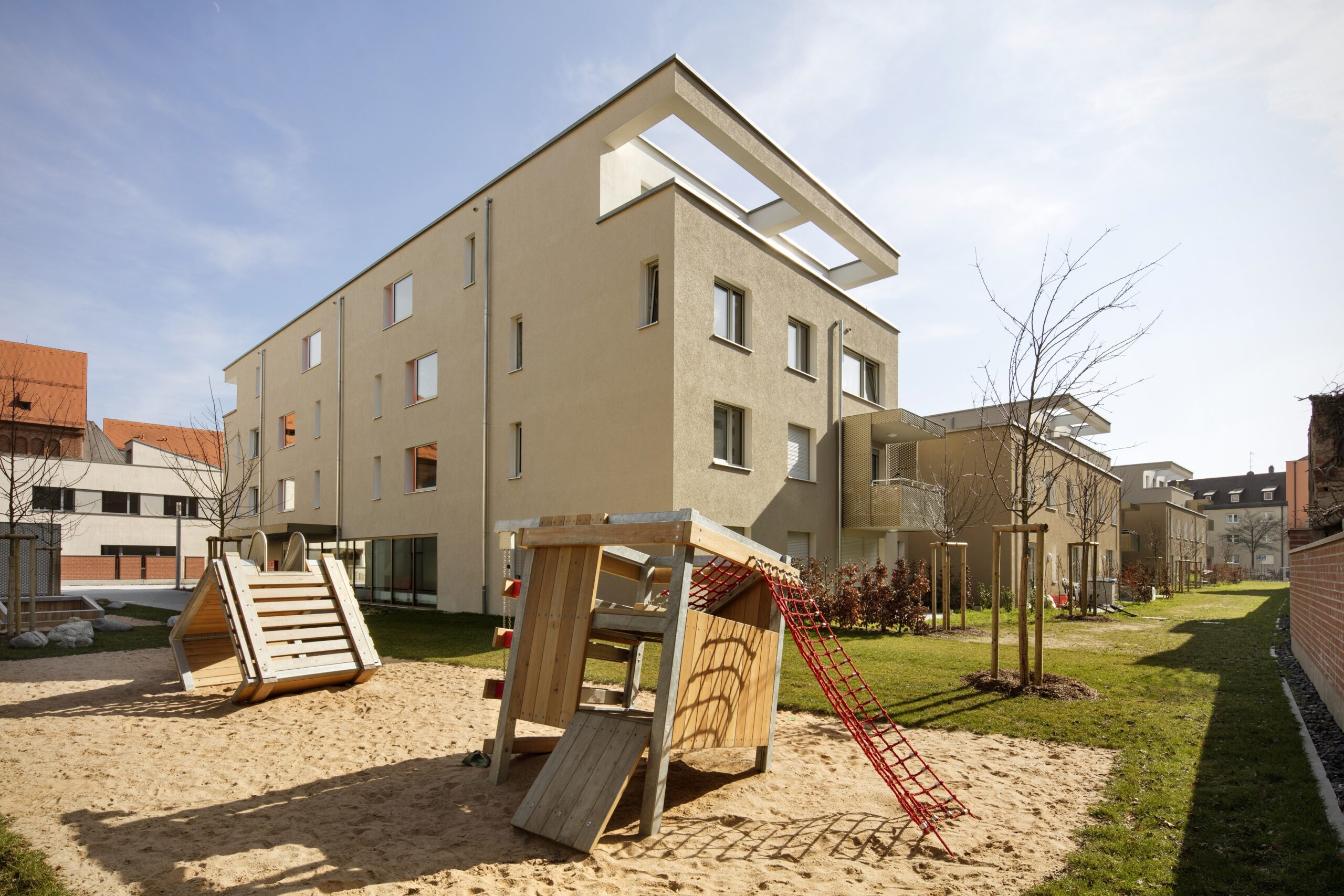 Der Apartmentkomplex in der Nähe der Hochschule Coburg besteht aus beigefarbenen Gebäuden inmitten üppiger Grünflächen und einem zentralen Spielplatz. Der Spielplatz bietet Holzkonstruktionen und Kletternetze auf einer Sandfläche, alles unter einem klaren, blauen Himmel.