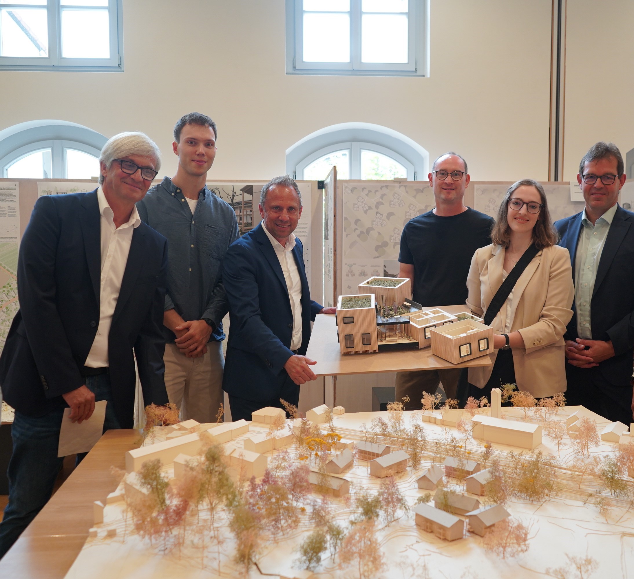 A group of people stands inside a room at Hochschule Coburg, gathered around a table with architectural models. They are smiling in their business or casual attire, with two individuals in the center proudly holding a model building. Behind them, display boards showcase various plans.