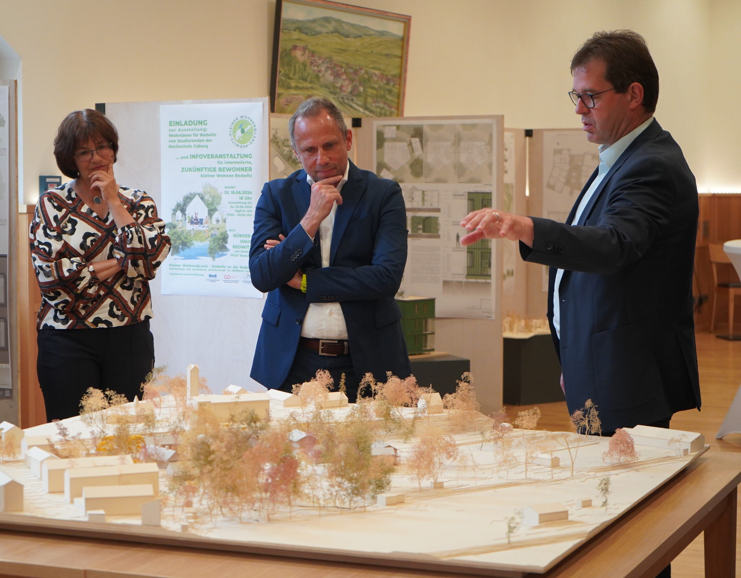 Three people are gathered around a 3D architectural model on a table at Hochschule Coburg. The man on the right gestures while speaking, as the other two listen attentively. Posters and plans are displayed in the background, showcasing creative projects from the university's talented students.