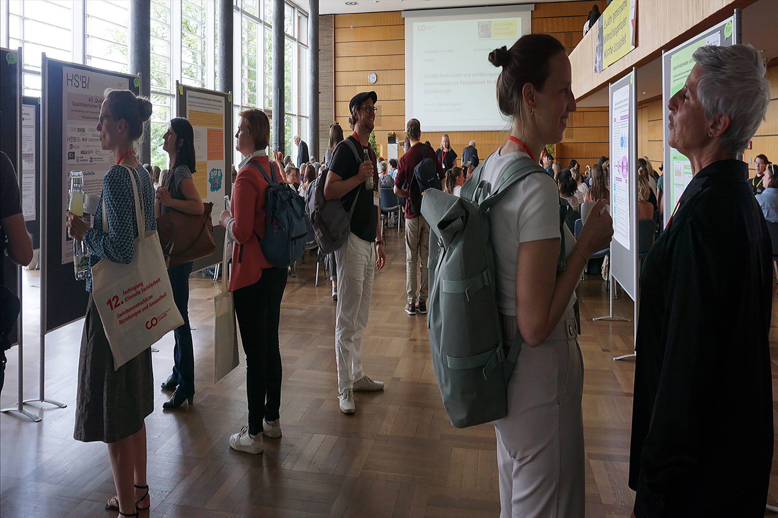 In einem hell erleuchteten Konferenzraum der Hochschule Coburg treffen sich die Leute und diskutieren an Forschungspostern. Einige tragen Rucksäcke und sind leger gekleidet, andere halten Getränke in der Hand. Große Fenster im Hintergrund lassen Tageslicht herein und schaffen eine einladende Atmosphäre für die Zusammenarbeit.