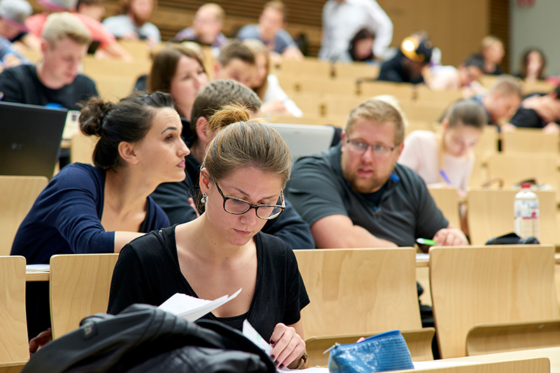 In einem Hörsaal der Hochschule Coburg sitzen die Studierenden in Reihen auf Holzbänken. Eine Frau im Vordergrund liest Aufsätze, während andere sich unterhalten oder Laptops benutzen. Im Raum herrscht reges Treiben, denn die Menschen sind in einen lebhaften Vortrag oder eine Diskussion vertieft.