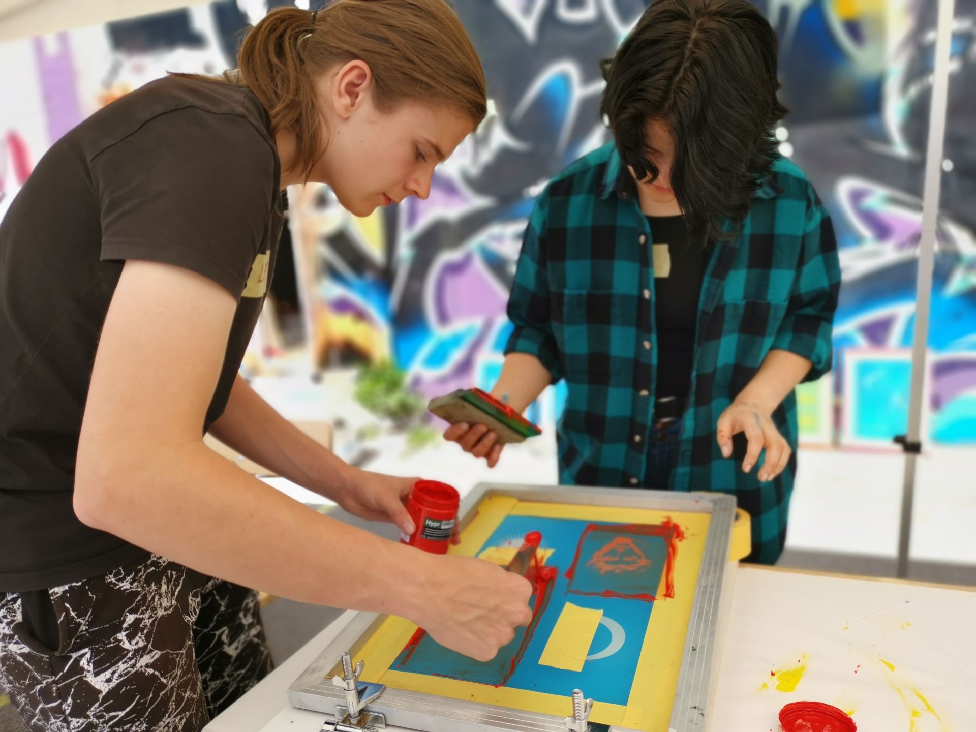 Zwei Personen an der Hochschule Coburg sind im Siebdruck tätig. Sie verwenden rote und grüne Tinte auf einer blauen Schablone. Sie konzentrieren sich intensiv auf ihre Arbeit, umgeben von lebendiger Graffiti-Kunst, die ihrem kreativen Prozess eine energetische Kulisse verleiht.