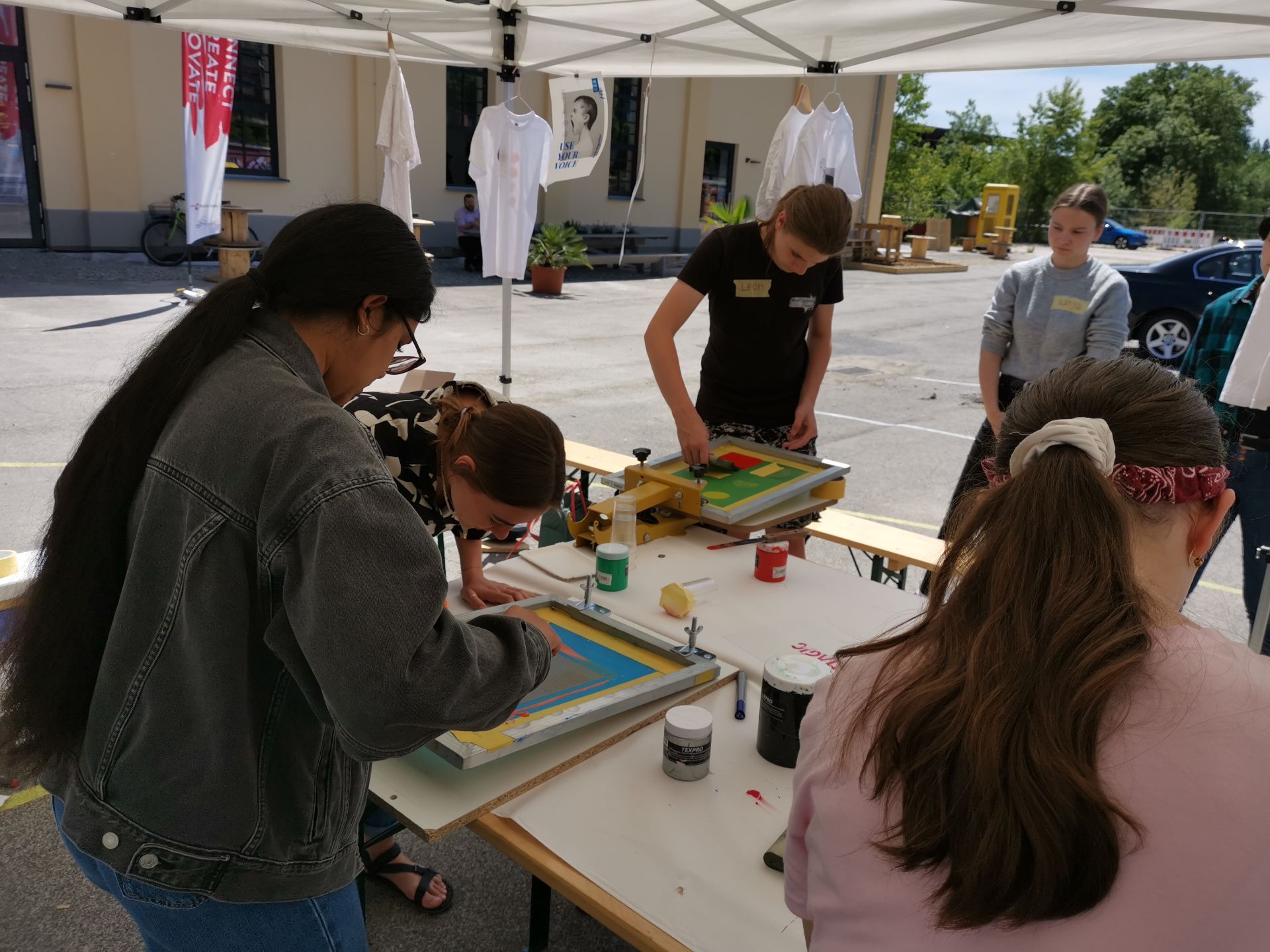 Eine Gruppe der Hochschule Coburg beschäftigt sich unter einem weißen Baldachin mit Siebdruck. Sie sind konzentriert und auf dem Tisch liegen verstreut Tinten und Rahmen. Im Hintergrund hängen ein paar T-Shirts und tragen zur kreativen Atmosphäre dieser sonnigen Umgebung im Freien bei.