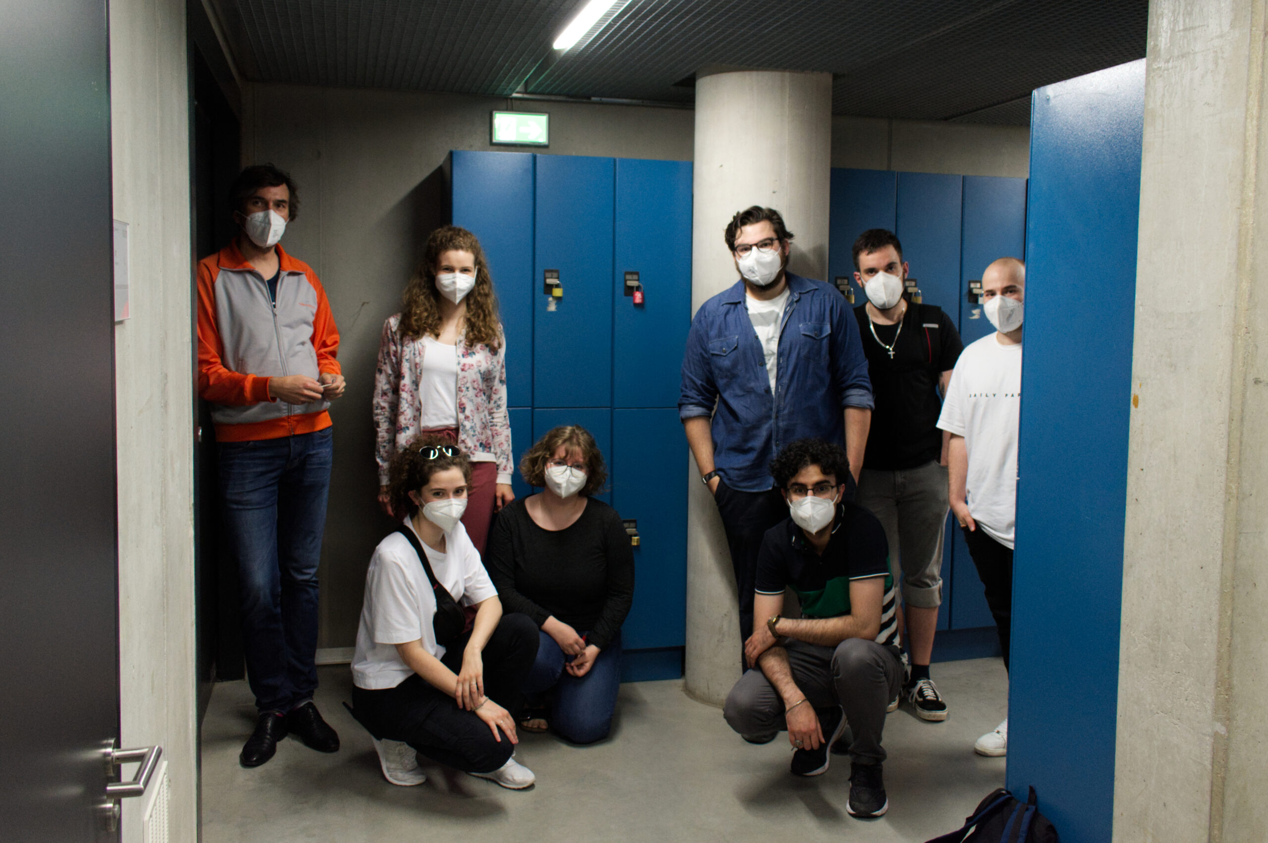 Eine Gruppe von acht maskierten Menschen steht in einem Flur der Hochschule Coburg, im Hintergrund sind blaue Schließfächer zu sehen. Einige sind leger gekleidet, andere knien, eingerahmt von Betonwänden und einer dunklen Decke.