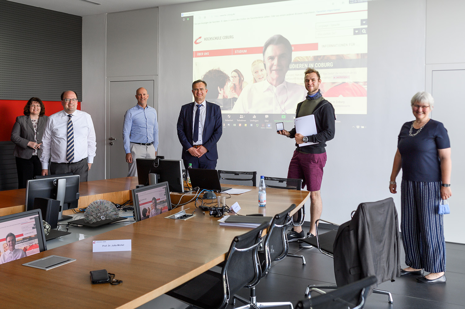 Eine Gruppe von sechs Personen steht in einem Konferenzraum der Hochschule Coburg. Sie sitzen mit etwas Abstand zueinander und lächeln, während hinter ihnen auf einer Projektionsfläche eine Website angezeigt wird. Auf dem Holztisch davor stehen mehrere Stühle und Computer.