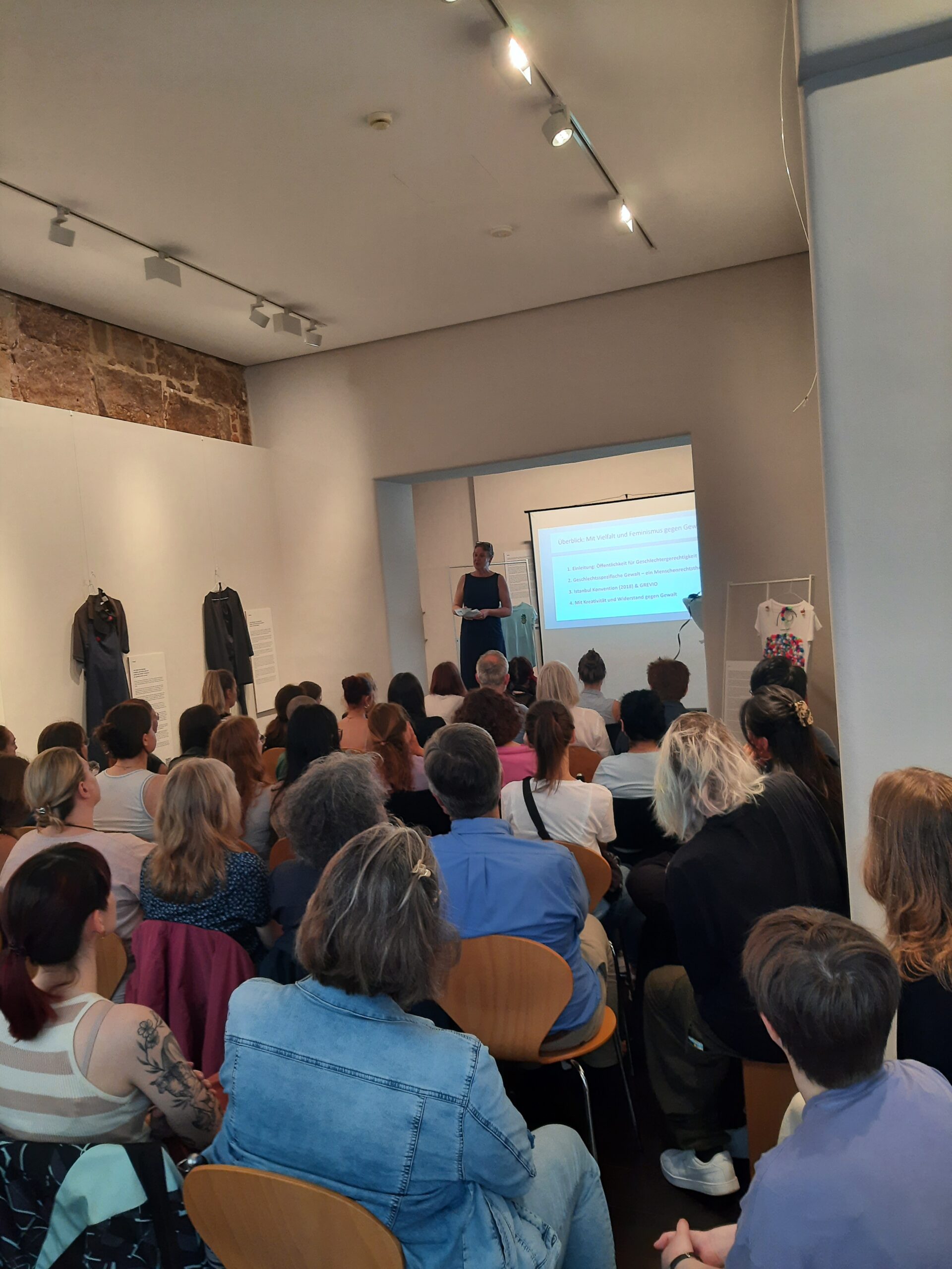 A speaker from Hochschule Coburg presents a slideshow to a seated audience in an art gallery. The room, adorned with white walls featuring artwork and clothing displays, creates a focused and engaging atmosphere as the attentive audience listens intently.