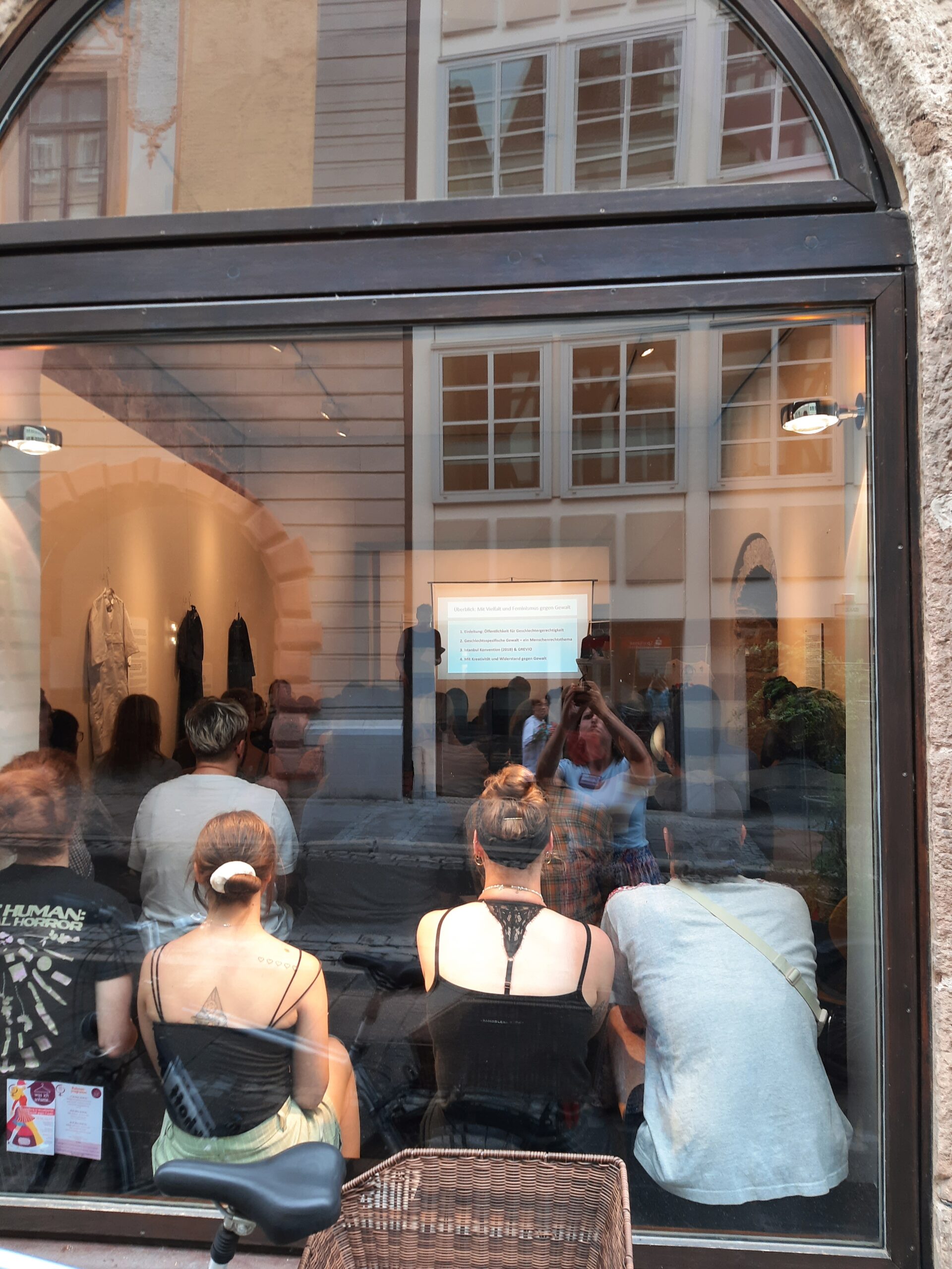 A group of people sits inside a room at Hochschule Coburg, viewed through a large window from the street. They appear to be attentively watching a presentation or screen at the front. A mix of casual outfits is visible, while the window reflects buildings from outside.