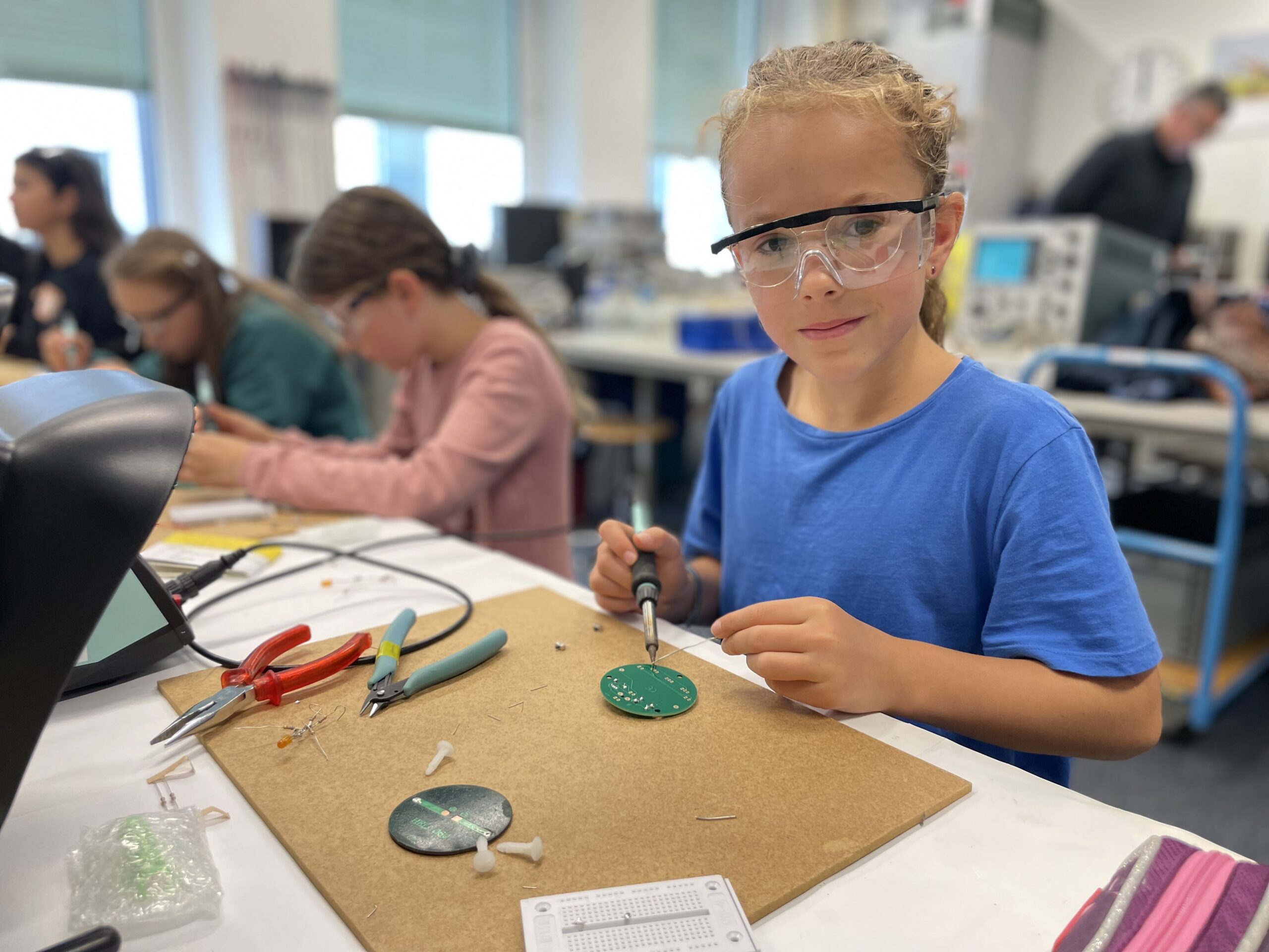 In einem Unterrichtsraum der Hochschule Coburg lötet ein junger Mensch mit Schutzbrille eine grüne Platine. Auf dem Tisch liegen verstreut Werkzeuge wie Zangen und Schneider, während im Hintergrund andere Studierende in ähnliche praktische Tätigkeiten vertieft sind.