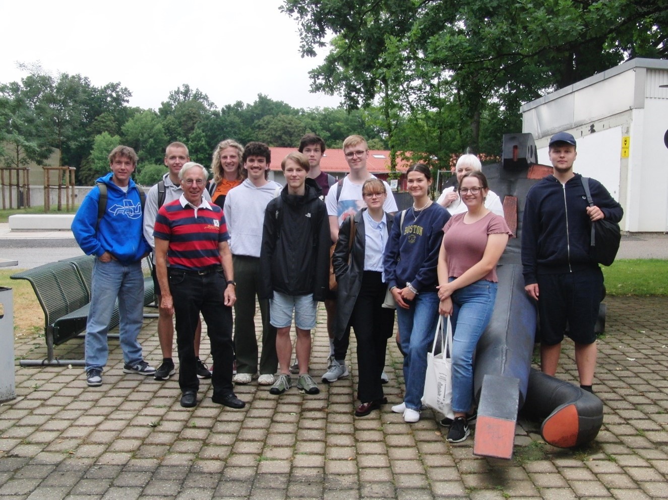 Eine Gruppe von dreizehn Personen, sowohl Männer als auch Frauen, steht entspannt draußen auf einer gepflasterten Fläche der Hochschule Coburg. Sie lächeln in die Kamera. Im Hintergrund sind Bäume und ein Gebäude zu sehen.