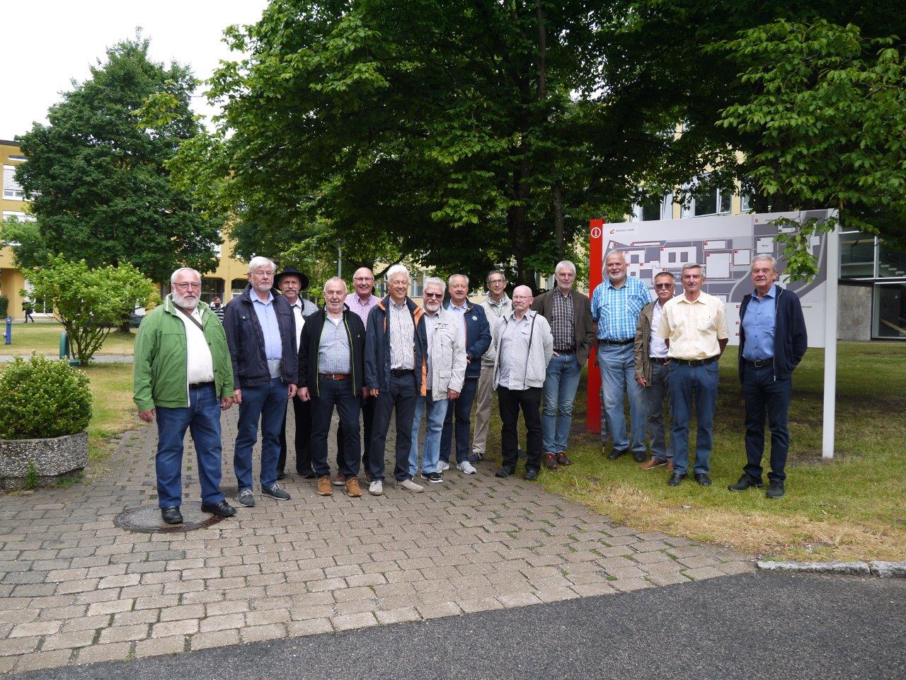 Eine Gruppe von Menschen steht draußen auf einem gepflasterten Weg an der Hochschule Coburg, im Hintergrund sind Bäume und ein Gebäude zu sehen. Sie posieren für ein Gruppenfoto, auf der rechten Seite sind etwas Grün und eine Karte oder ein Schild zu sehen.