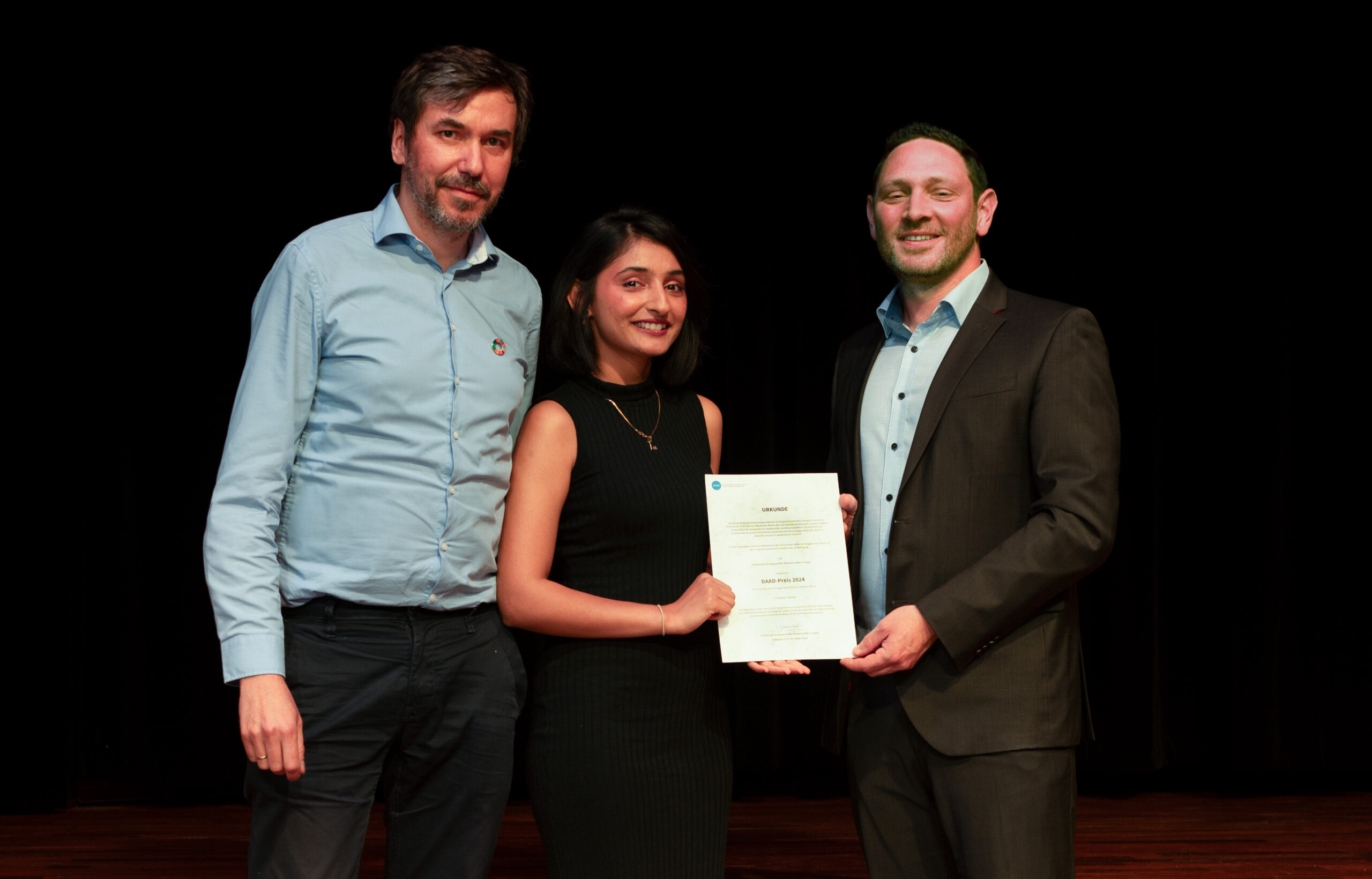 Three people stand together on a stage at Hochschule Coburg. The person in the center holds a certificate, smiling, while the other two people stand on either side, also smiling. The background is a dark curtain.