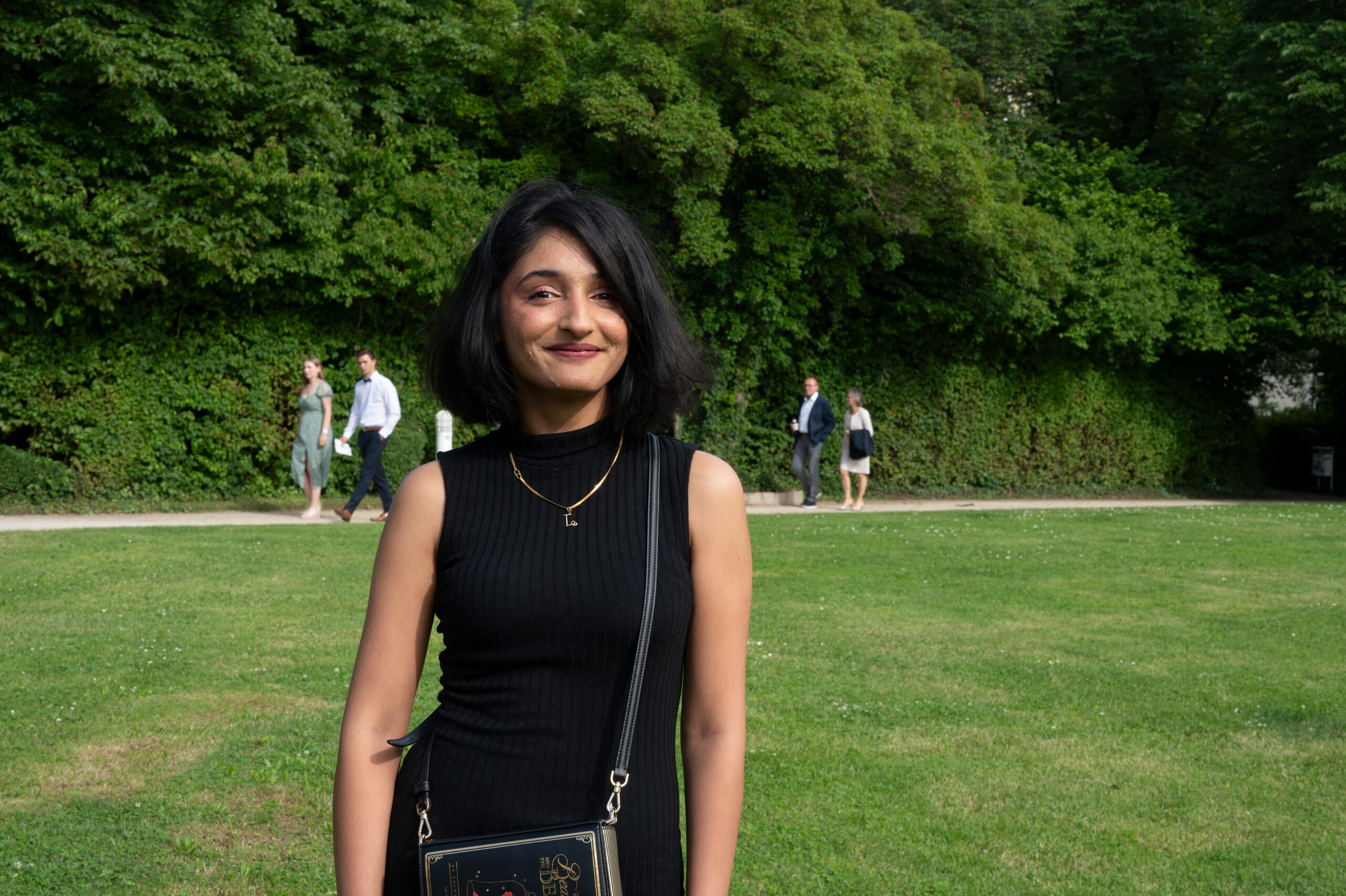 A woman in a black dress stands smiling in a park near Hochschule Coburg, with lush green trees in the background. A few people are walking on a path behind her.