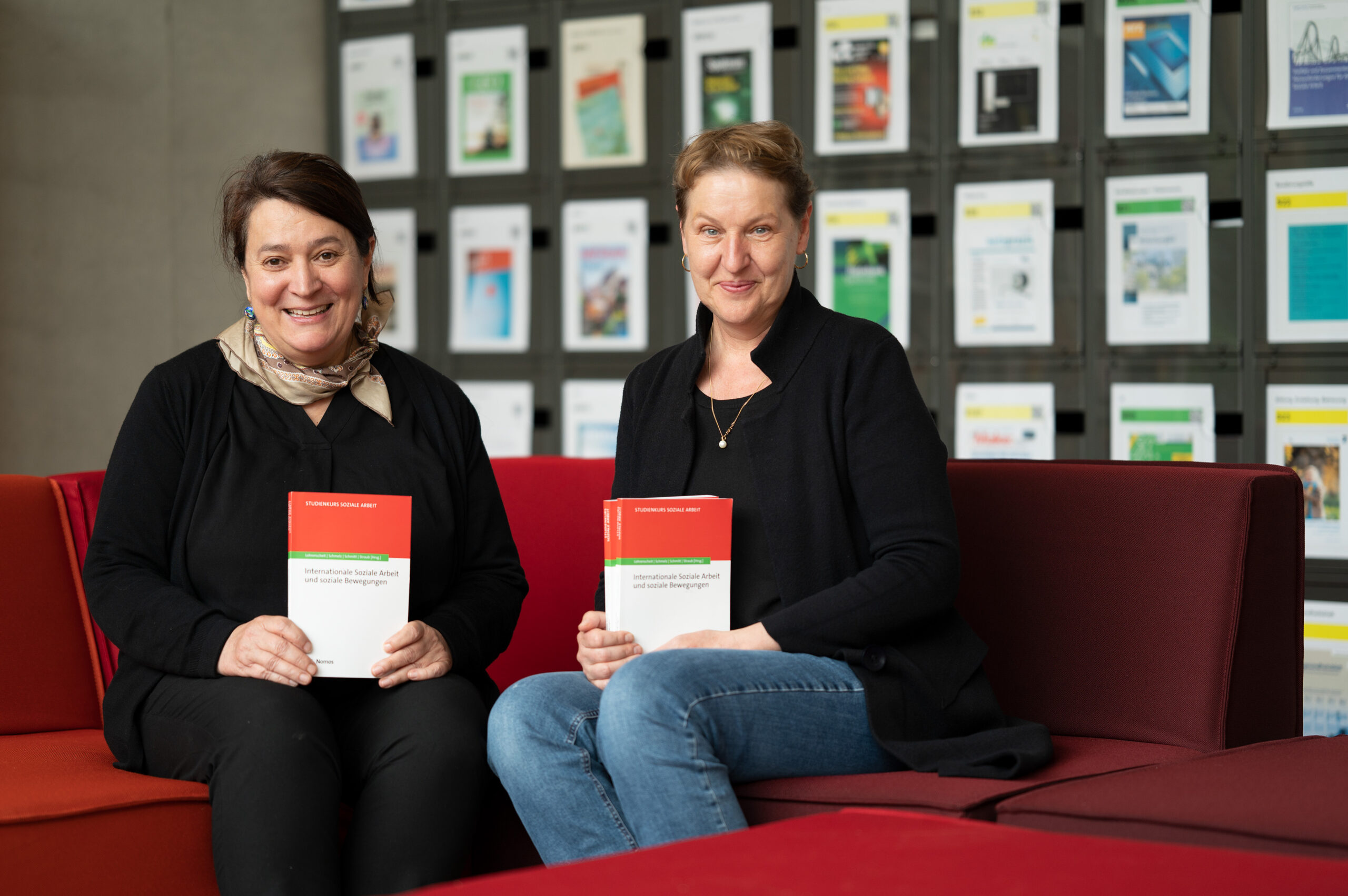 Zwei Frauen sitzen auf einem roten Sofa in der Hochschule Coburg und halten Bücher mit dem Titel „Communications Engineering Desk Reference“ in den Händen. Sie stehen vor einer Wand, die mit verschiedenen Publikationen geschmückt ist, wodurch eine akademische Atmosphäre entsteht, die an eine Bibliothek erinnert.