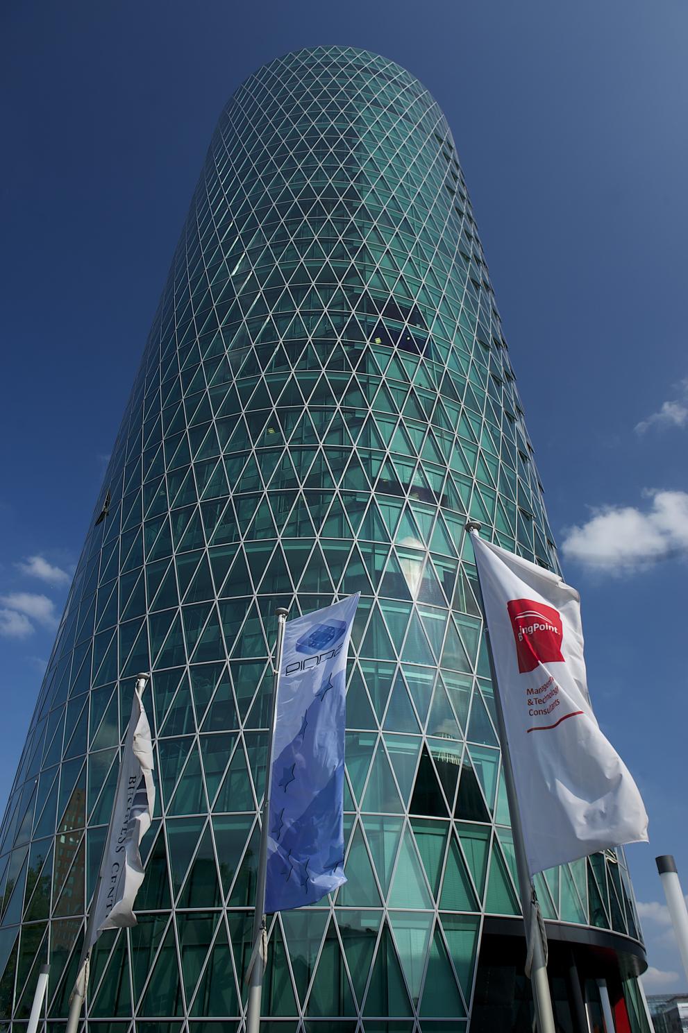 A tall, cylindrical glass skyscraper with a diamond-patterned facade stands proudly, reminiscent of the innovative design celebrated at Hochschule Coburg. White flags wave gracefully in the foreground against a clear blue sky.