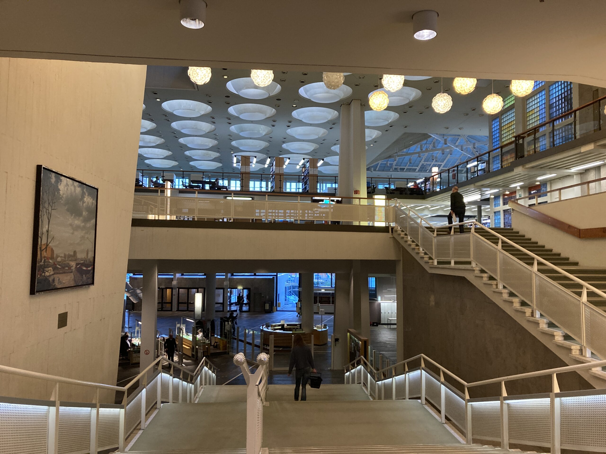 Weite Aufnahme eines geräumigen, modernen Bibliotheksinnenraums der Hochschule Coburg mit mehreren Stockwerken. Große runde Lampen hängen von der Decke und Besucher gehen die weißen Treppen hinauf und hinunter. An einer Wand links hängt ein Gemälde, das dieser akademischen Oase einen Hauch von Eleganz verleiht.