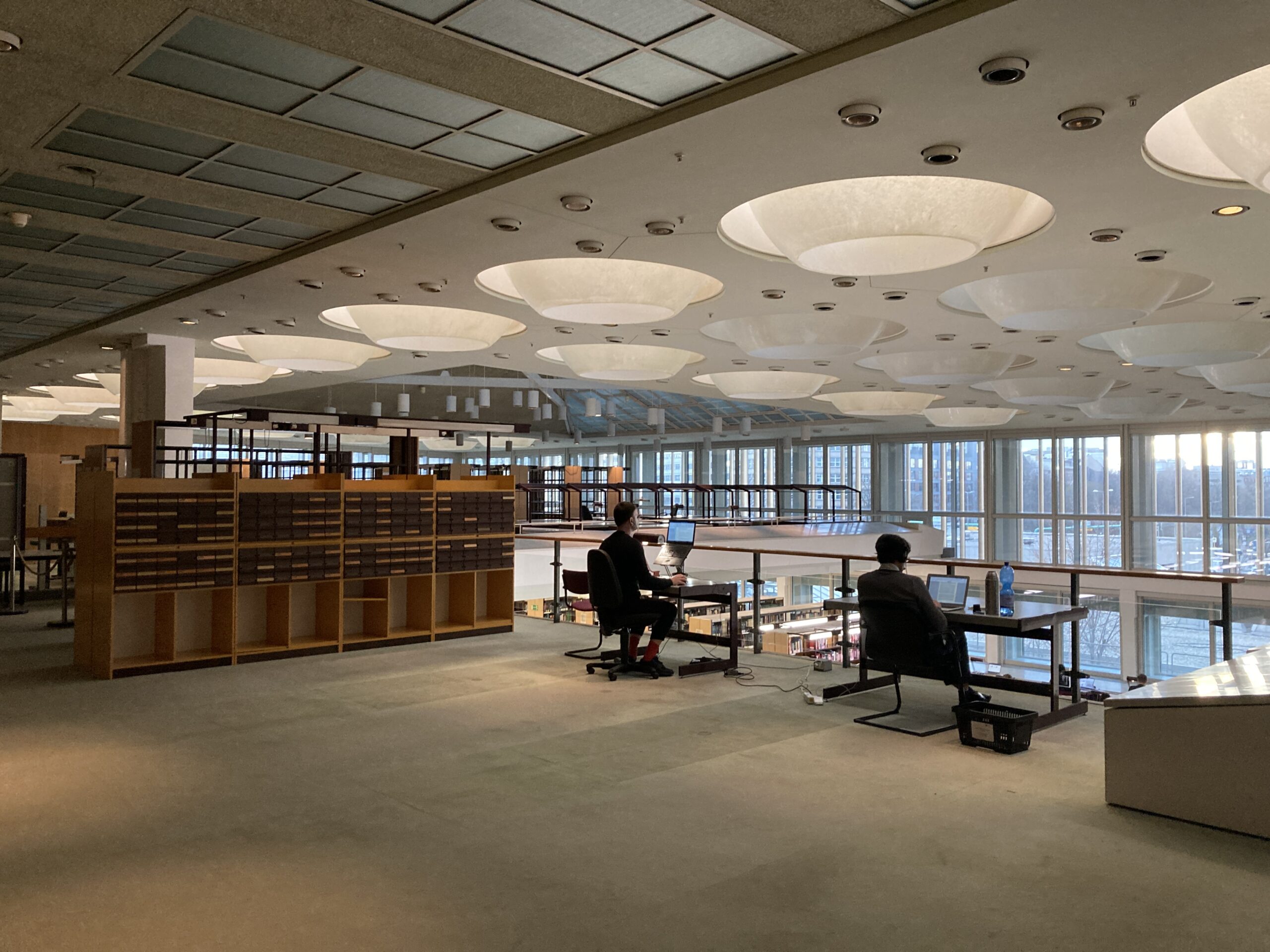 Großer Büroraum mit hohen Decken und geometrischen Leuchten an der Hochschule Coburg. Zwei Personen sitzen an Schreibtischen neben einem großen Fenster mit Blick auf die Stadtlandschaft. Daneben steht ein beleuchtetes Regal, und das natürliche Licht schafft eine geräumige, moderne Atmosphäre.