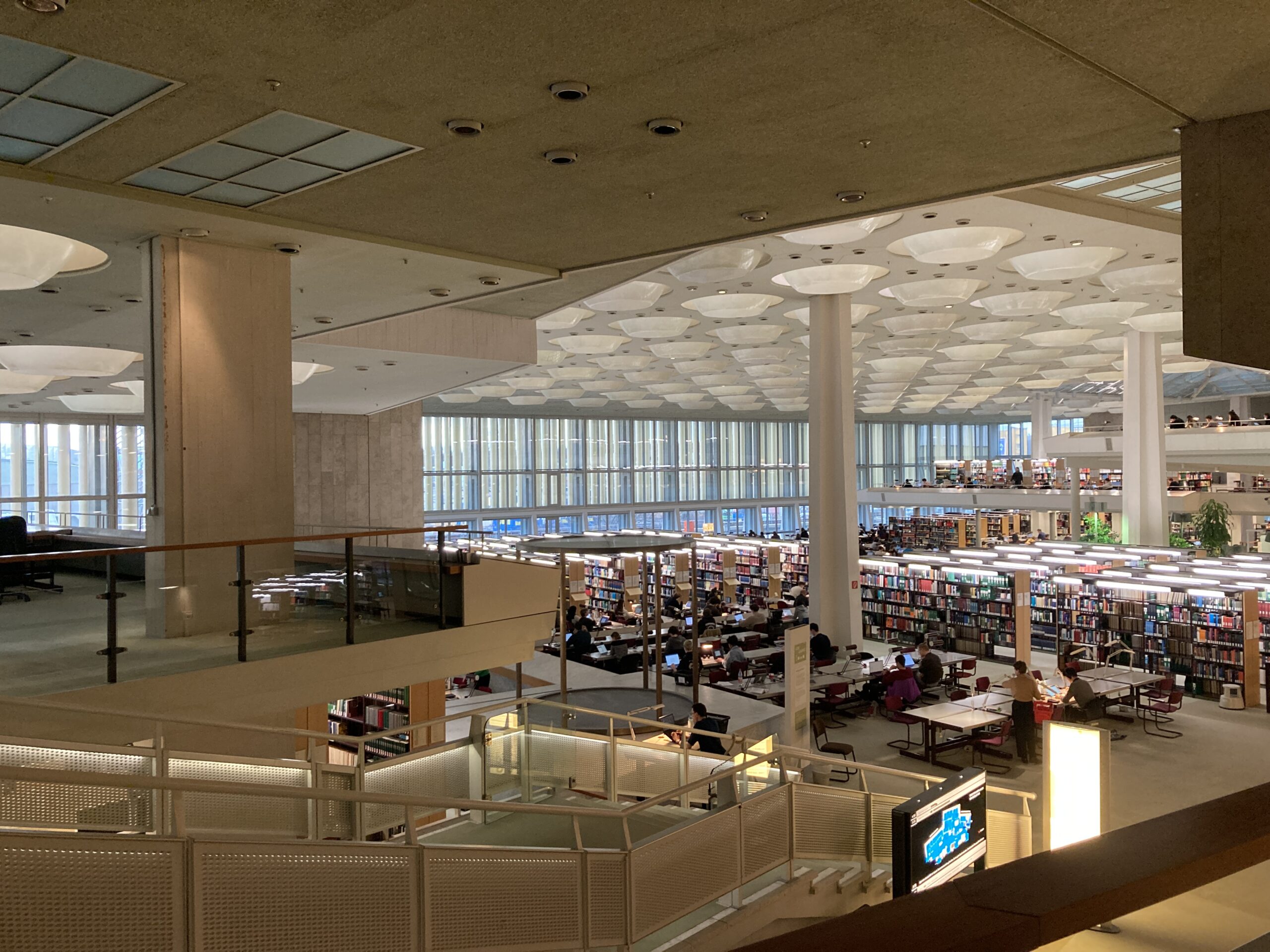 Der geräumige Bibliotheksinnenraum der Hochschule Coburg besticht durch hohe Decken und große Fenster. Reihen von Bücherregalen füllen die untere Ebene, wo Menschen an Tischen sitzen und lesen und arbeiten. Die obere Ebene verfügt über einen offenen Gang mit modernem Design, der eine perfekte Mischung aus Tradition und Innovation verkörpert.