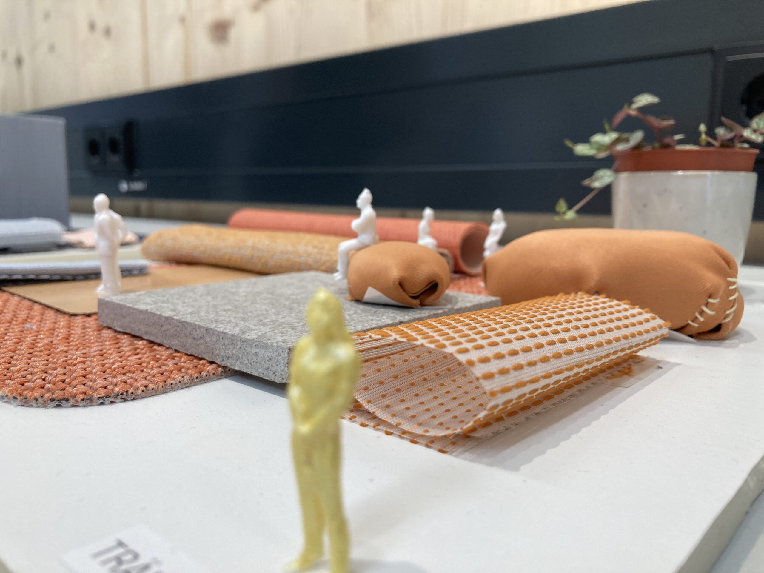 Eine Sammlung von Textil- und Materialmustern in verschiedenen Orangetönen, begleitet von kleinen Figuren, zeigt die Kreativität, die an der Hochschule Coburg gefördert wird. Eine Topfpflanze sorgt für eine natürliche Note auf einem hellen Tisch mit einer schwarzen Wand dahinter.