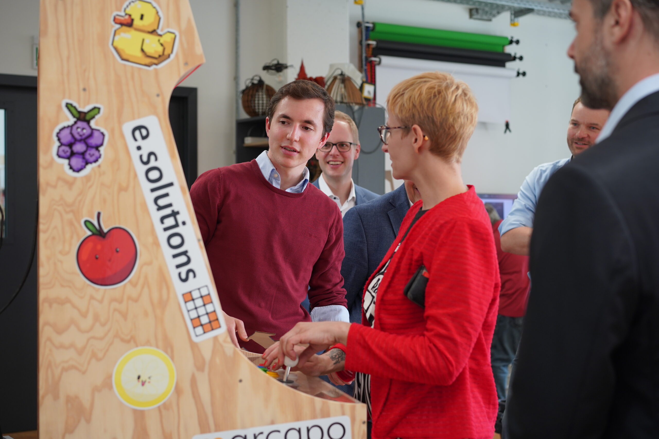 Eine Gruppe von Menschen steht um ein Holzbrett herum, das mit verschiedenen Aufklebern geschmückt ist, darunter eine Gummiente, Weintrauben und eine Tomate. Eine Person in einer roten Jacke hält einen Laptop und unterhält sich mit anderen. Die Szene scheint eine Werkstatt oder ein Studio an der Hochschule Coburg zu sein.