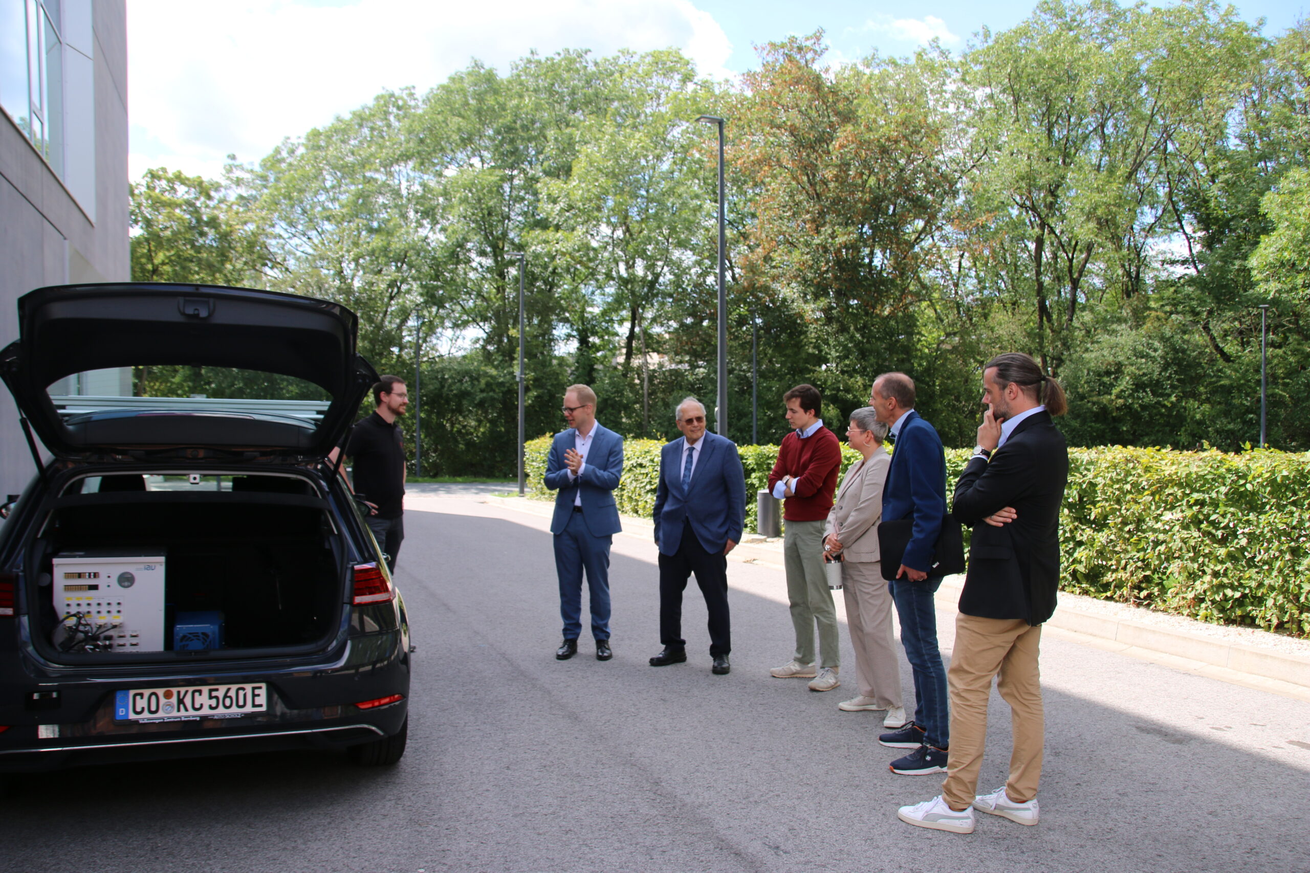 Eine Gruppe von Menschen steht draußen neben einem schwarzen Auto mit offenem Kofferraum voller elektronischer Geräte, anscheinend ein Projekt von Studenten der Hochschule Coburg. Bäume und Büsche rahmen die Szene unter einem teilweise bewölkten Himmel ein, während sie sich angeregt über das Auto unterhalten.