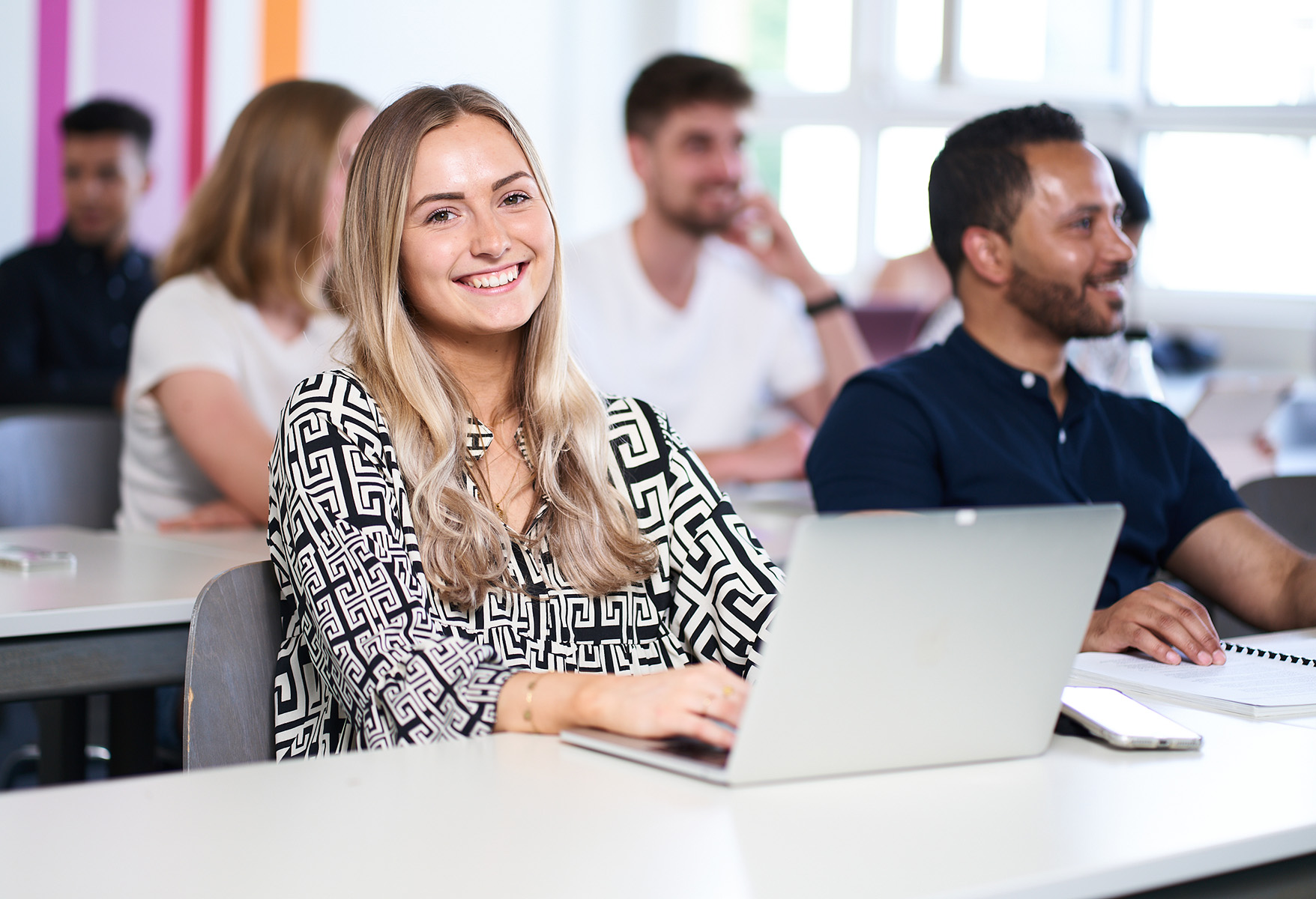 Eine Frau mit langen blonden Haaren lächelt in einem hellen, gut beleuchteten Hörsaal der Hochschule Coburg in die Kamera. Vor ihr liegt ihr aufgeklappter Laptop. Im Hintergrund sind mehrere andere Studierende in eine lebhafte Vorlesung oder Diskussion vertieft.