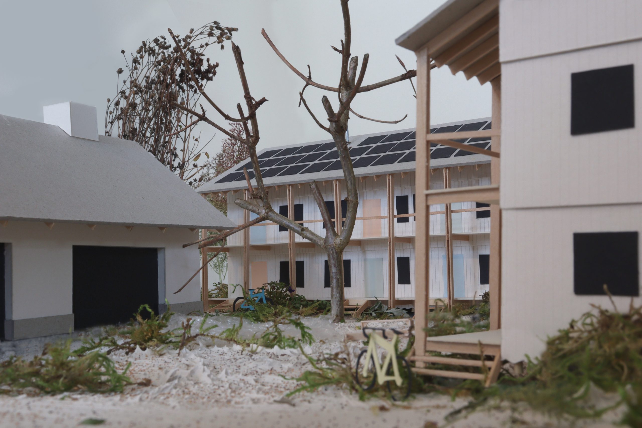 A detailed architectural model showcases a courtyard scene at Hochschule Coburg, with a tree at the center. Surrounding buildings boast modern design featuring solar panels, flat roofs, and wooden structures. Small plants and a bicycle add charm to this sustainable academic setting.