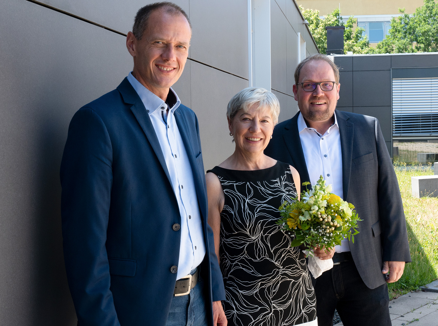 Drei formell gekleidete Menschen stehen im Freien vor dem modernen Gebäude der Hochschule Coburg. Die Frau in der Mitte hält einen Strauß gelber und weißer Blumen in der Hand, im Hintergrund ist üppiges Grün zu sehen.