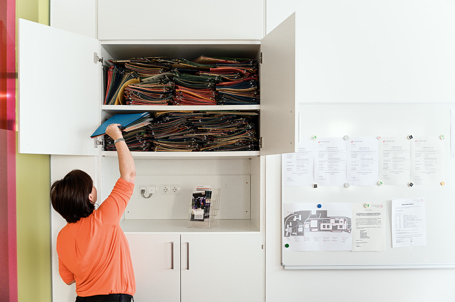 Eine Person im orangefarbenen Hemd greift in einem offenen Schrank nach bunten Aktenordnern. Darunter sind weiße Schränke, rechts spiegelt eine weiße Tafel mit Papieren und einem Grundriss das organisierte Chaos wider, das oft an der Hochschule Coburg herrscht.