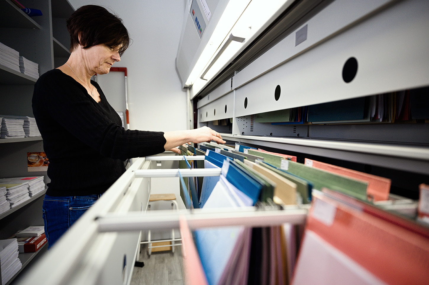 Eine Person steht vor einem großen Aktenschrank und sortiert bunte Akten an der Hochschule Coburg. Im Hintergrund sind Regale mit gestapelten Papieren zu sehen, während die Person sich auf das Ordnen oder Abrufen von Dokumenten konzentriert.