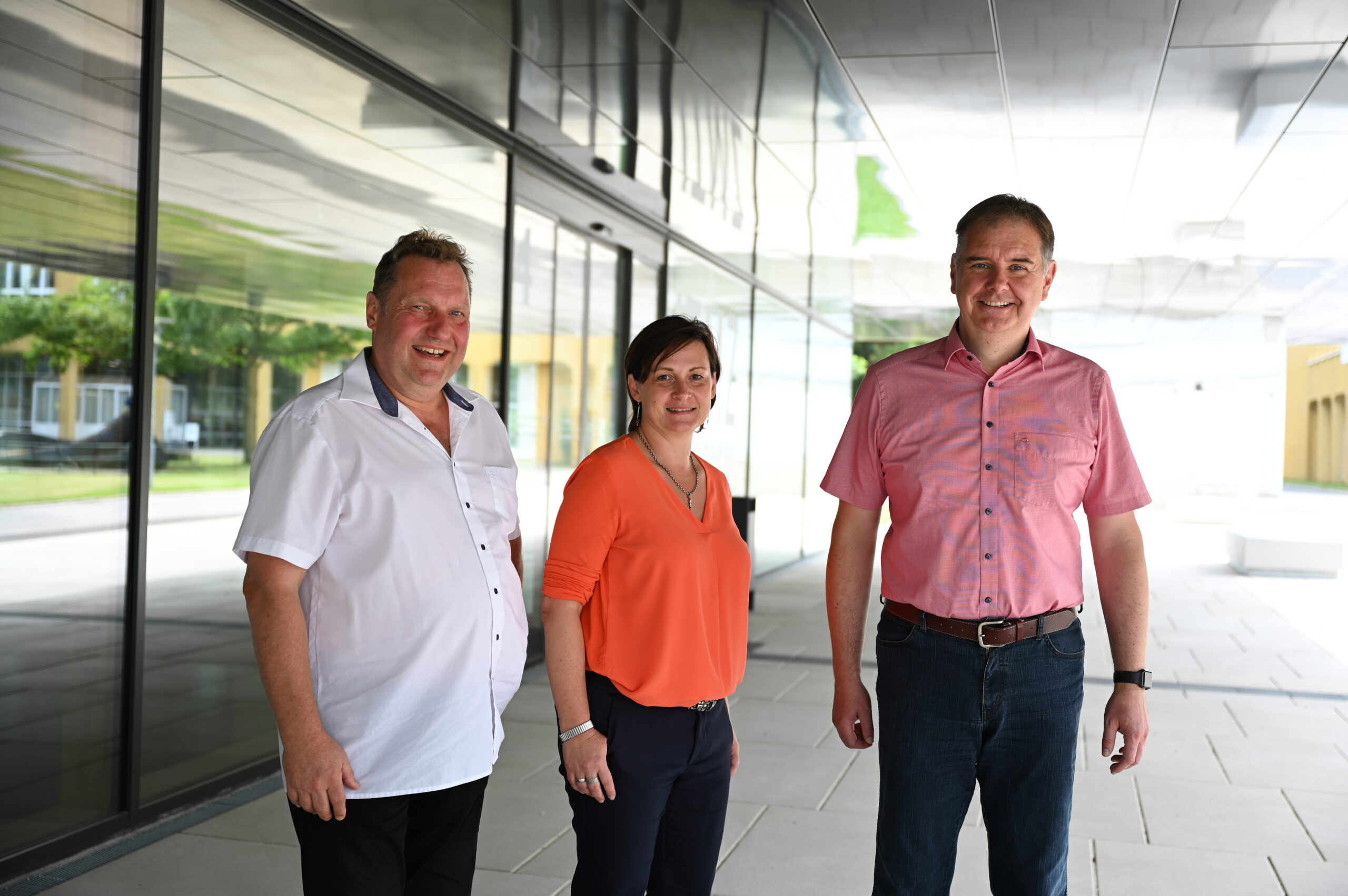 Drei Personen stehen vor dem modernen Gebäude der Hochschule Coburg mit seinen spiegelnden Glaswänden. Die Gruppe besteht aus zwei Männern und einer Frau, alle lächeln. Der Mann links trägt ein weißes Hemd, die Frau ein orangefarbenes Oberteil und der Mann rechts ein rosa Hemd.