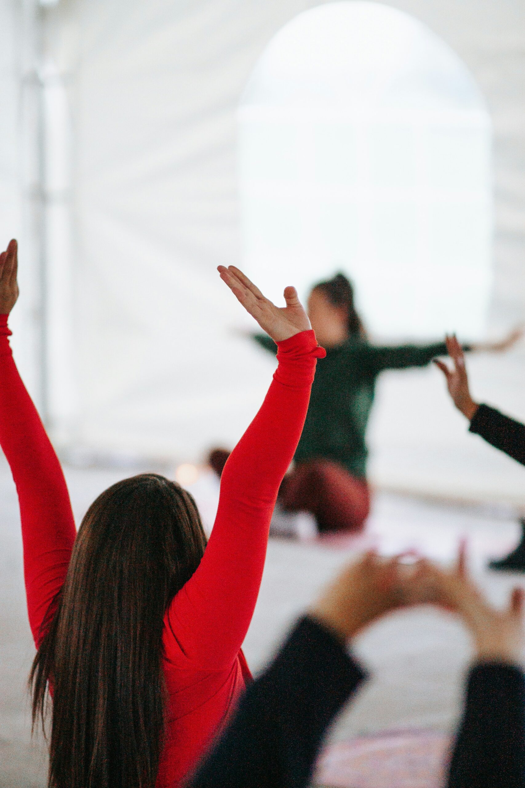 Eine Gruppe von Menschen praktiziert Yoga in Innenräumen der Hochschule Coburg. Eine Person im Vordergrund trägt ein rotes Hemd und hat die Arme erhoben, während andere im Hintergrund verschwommen im Schneidersitz mit ausgestreckten Armen sitzen. Die Umgebung ist hell und natürlich.