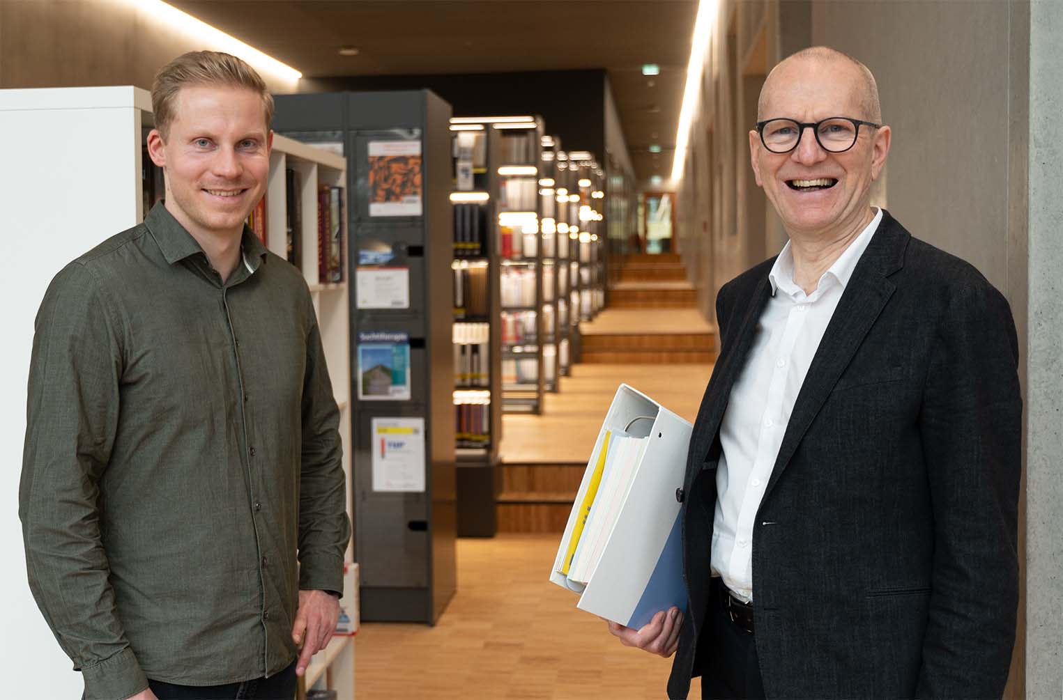 Zwei Männer lächeln in einer modernen Bibliothek der Hochschule Coburg. Einer hält Ordner in der Hand und trägt Brille und Anzug, während der andere ein legeres Hemd trägt. Sie stehen in einem mit Bücherregalen gesäumten Korridor und verkörpern die Mischung aus Professionalität und entspanntem Lernen an dieser renommierten Institution.