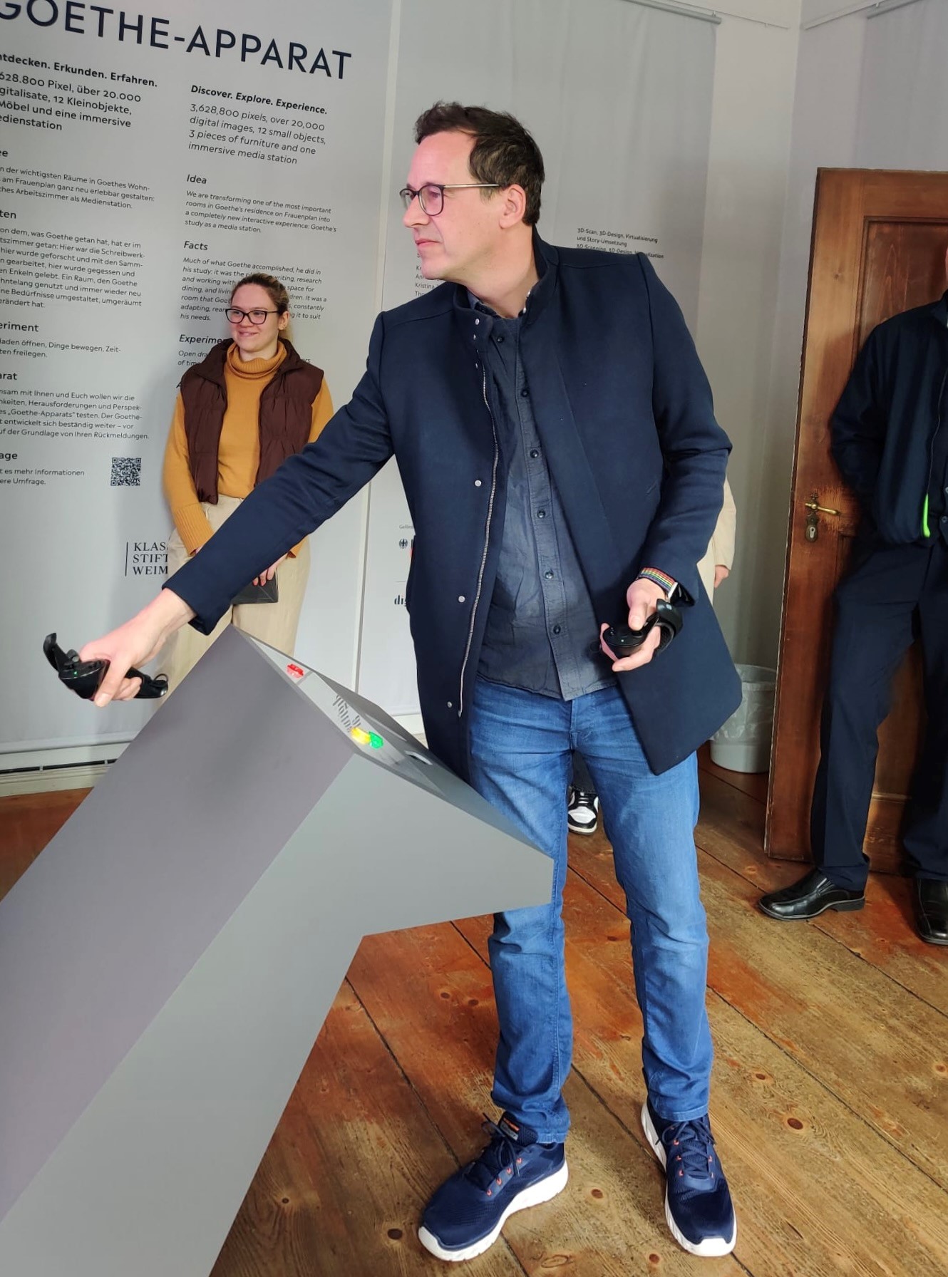 A man wearing a dark blazer and glasses interacts with a display at an exhibit, likely part of the Hochschule Coburg showcase. A woman stands in the background, smiling. Both are in a room with wooden floors and informational posters on the wall.