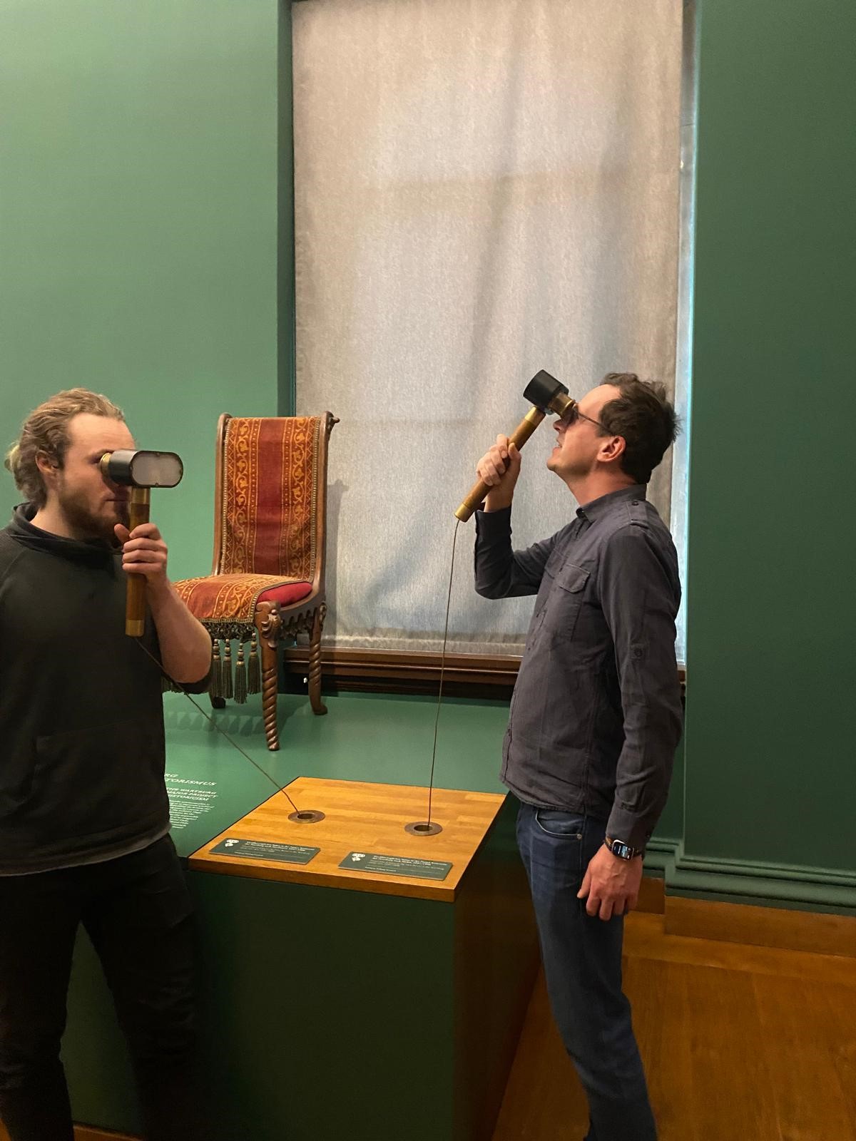 Two men humorously pose with their heads tilted back, pressing oversized rubber mallets to their foreheads at a Hochschule Coburg exhibit. In the background, a minimalist display showcases a chair mounted on the wall with informative plaques below.