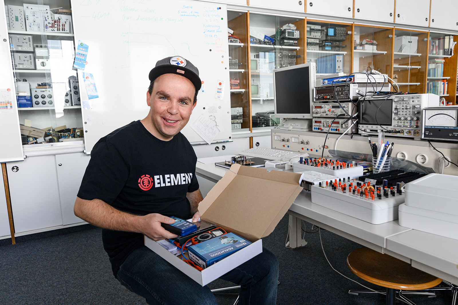 Eine Person in einem Labor der Hochschule Coburg lächelt, während sie einen offenen Karton mit elektronischen Bauteilen in der Hand hält. Der Raum ist mit Regalen, einem Whiteboard und verschiedenen elektronischen Geräten und Werkzeugen auf einem Schreibtisch ausgestattet.