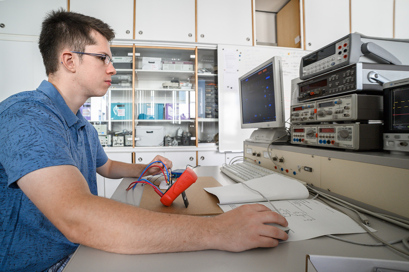 Eine Person im blauen Hemd sitzt an einem Schreibtisch in einem Labor der Hochschule Coburg, umgeben von elektronischen Geräten mit mehreren Bildschirmen und Messgeräten. Sie hält ein rotes Gerät mit Kabeln in der Hand und analysiert dessen Messwerte auf einem Monitor. Auf dem Schreibtisch liegen Papiere ausgebreitet, die auf detaillierte Forschungsarbeit hindeuten.