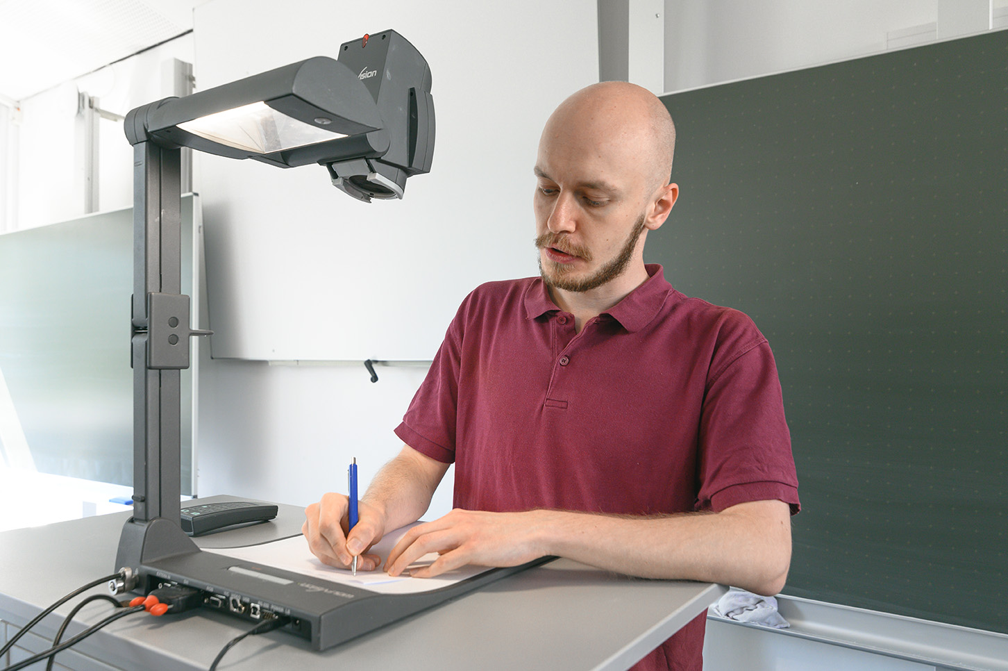 Ein Mann in einem kastanienbraunen Hemd von der Hochschule Coburg benutzt einen Overheadprojektor, um Notizen auf ein Blatt Papier zu schreiben. Er steht in einem hellen Klassenzimmer mit einer grünen Tafel im Hintergrund.