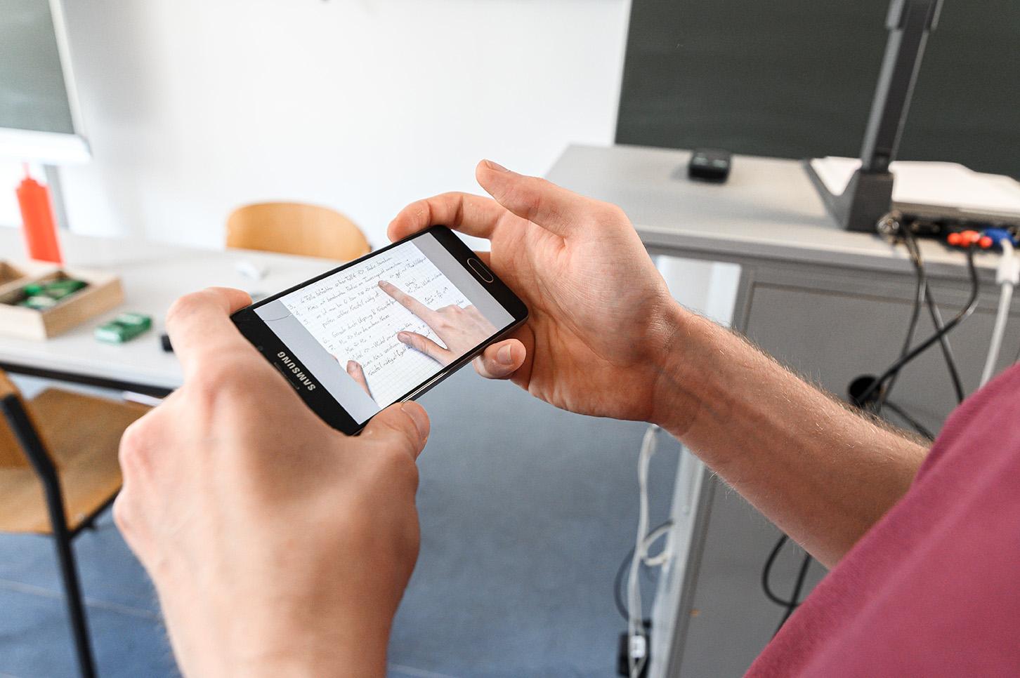 Eine Person an der Hochschule Coburg hält ein Samsung-Smartphone waagerecht und macht ein Foto von handschriftlichen Notizen. Im Hintergrund sind im Klassenzimmer ein Tisch, ein Stuhl und einige elektronische Geräte zu sehen.