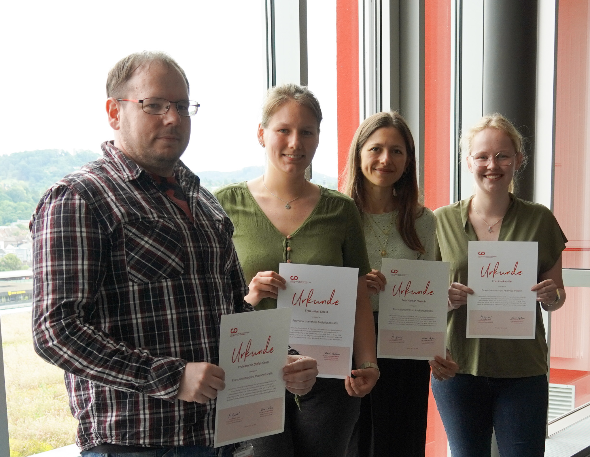 Four people stand indoors at Hochschule Coburg, proudly holding certificates and smiling. They are near a large window with a view of lush greenery outside.