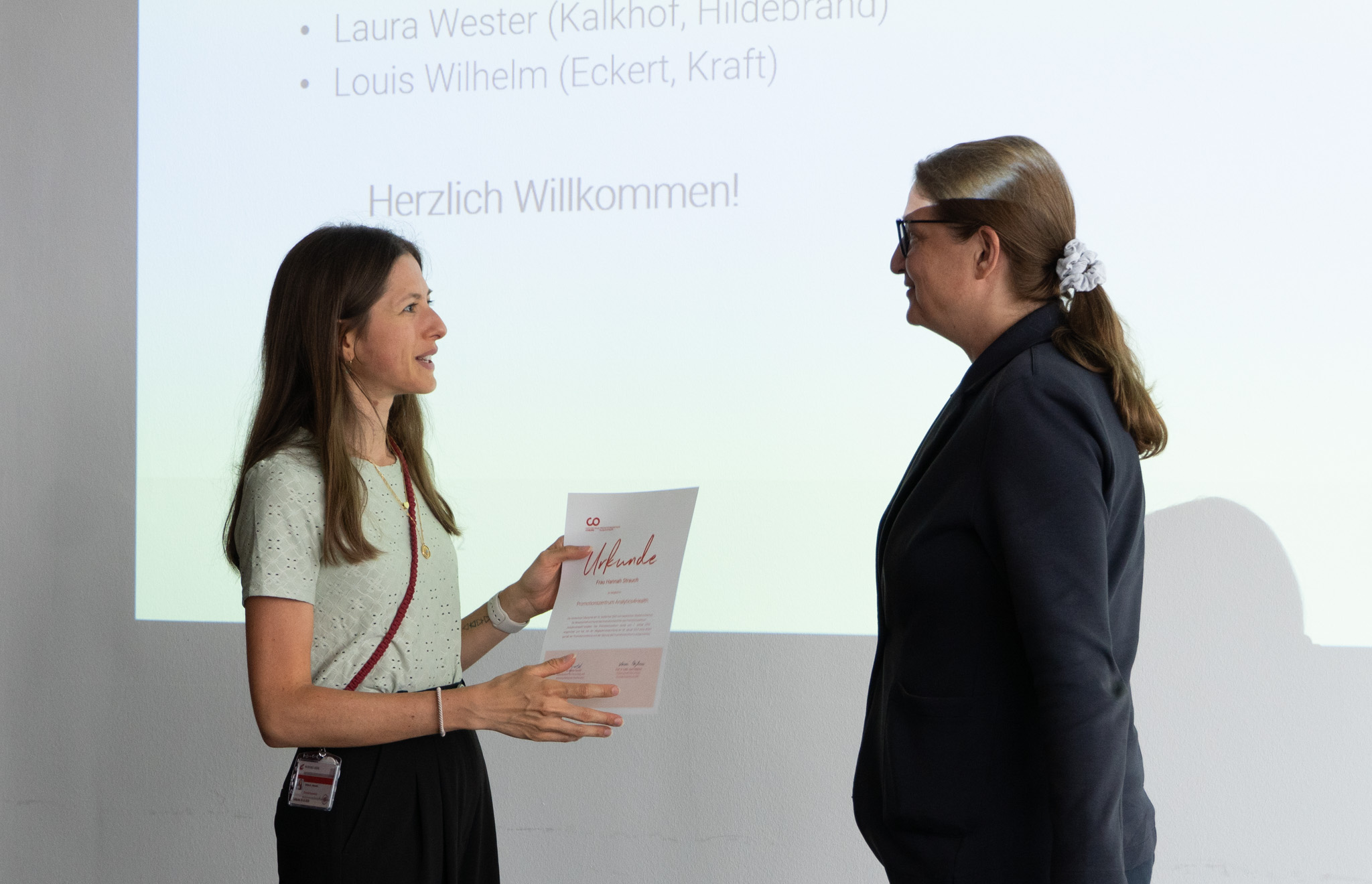 Two women stand facing each other in front of a presentation screen at Hochschule Coburg. One is handing a certificate to the other. The projected slide includes the words "Herzlich Willkommen!" and names like Laura Wester and Louis Wilhelm.