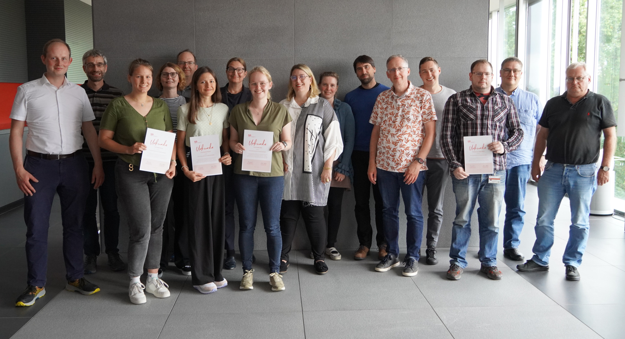 A group of 15 people stands in a room at Hochschule Coburg, some proudly holding certificates. They are dressed casually and smiling at the camera. The space features gray walls and large windows, creating a bright and welcoming atmosphere.